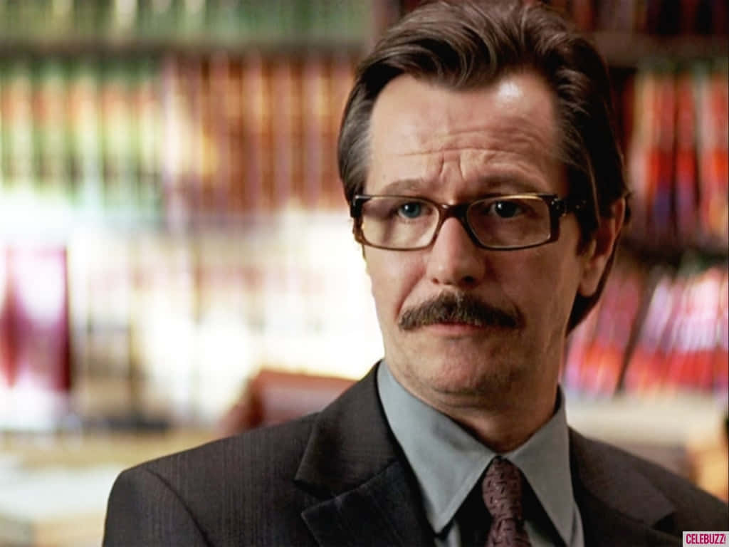 Commissioner Gordon 桌布 壁紙 — 一位戴眼鏡、留鬍子、身穿西裝的男子站在一個模糊的書架前，書架上擺滿了色彩繽紛的書籍。