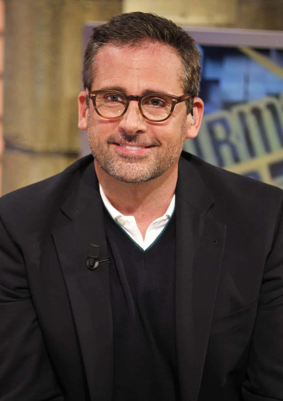 steve carell 桌布 壁紙 — 一位戴眼鏡、留鬍子的男士穿著黑色西裝和毛衣背心，溫暖地微笑。