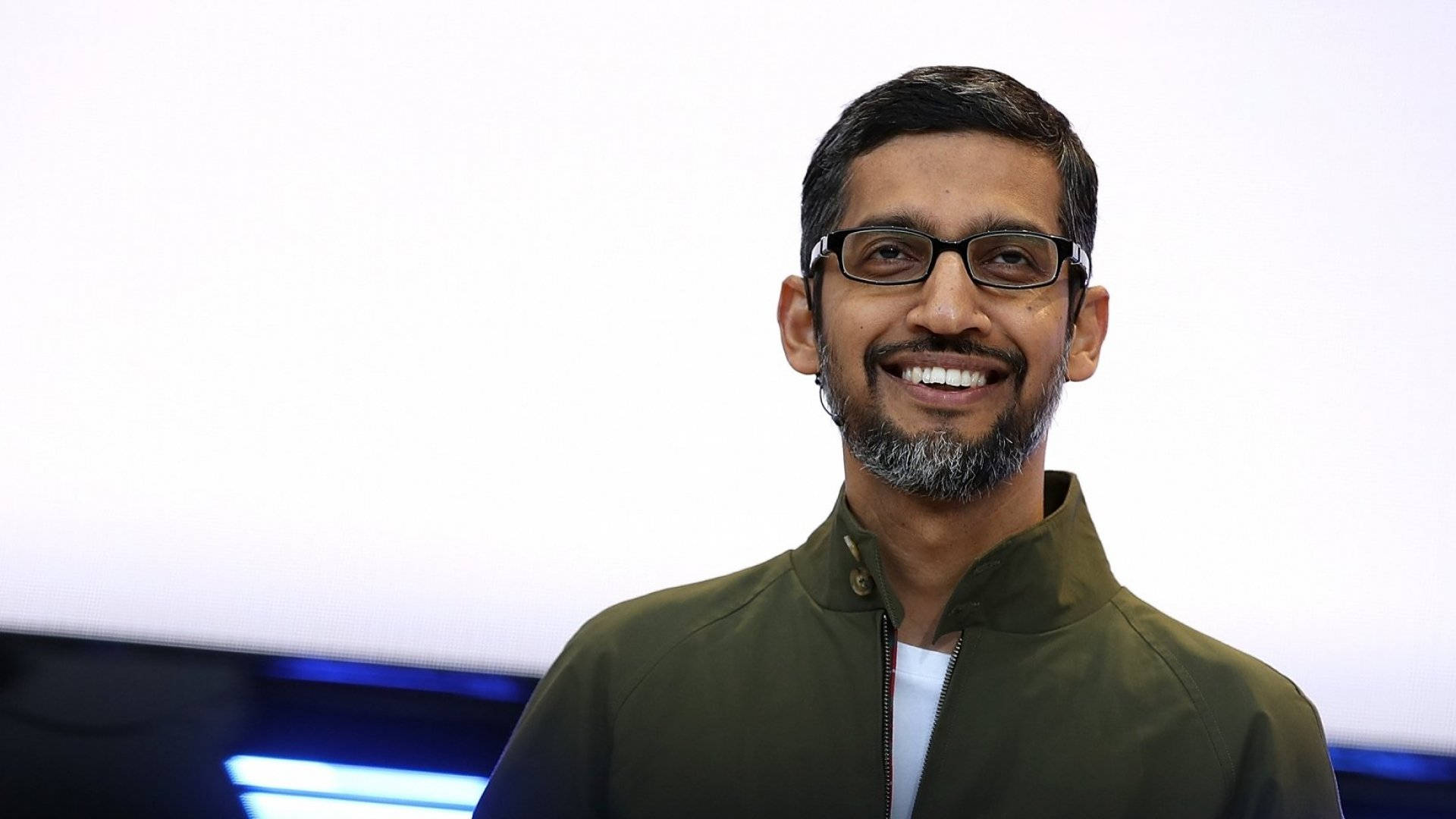 google 桌布 壁紙 — 一位戴眼鏡、留鬍鬚、面帶微笑的男子，身穿橄欖色外套，背景是明亮的白色，帶有藍色燈光點綴。
