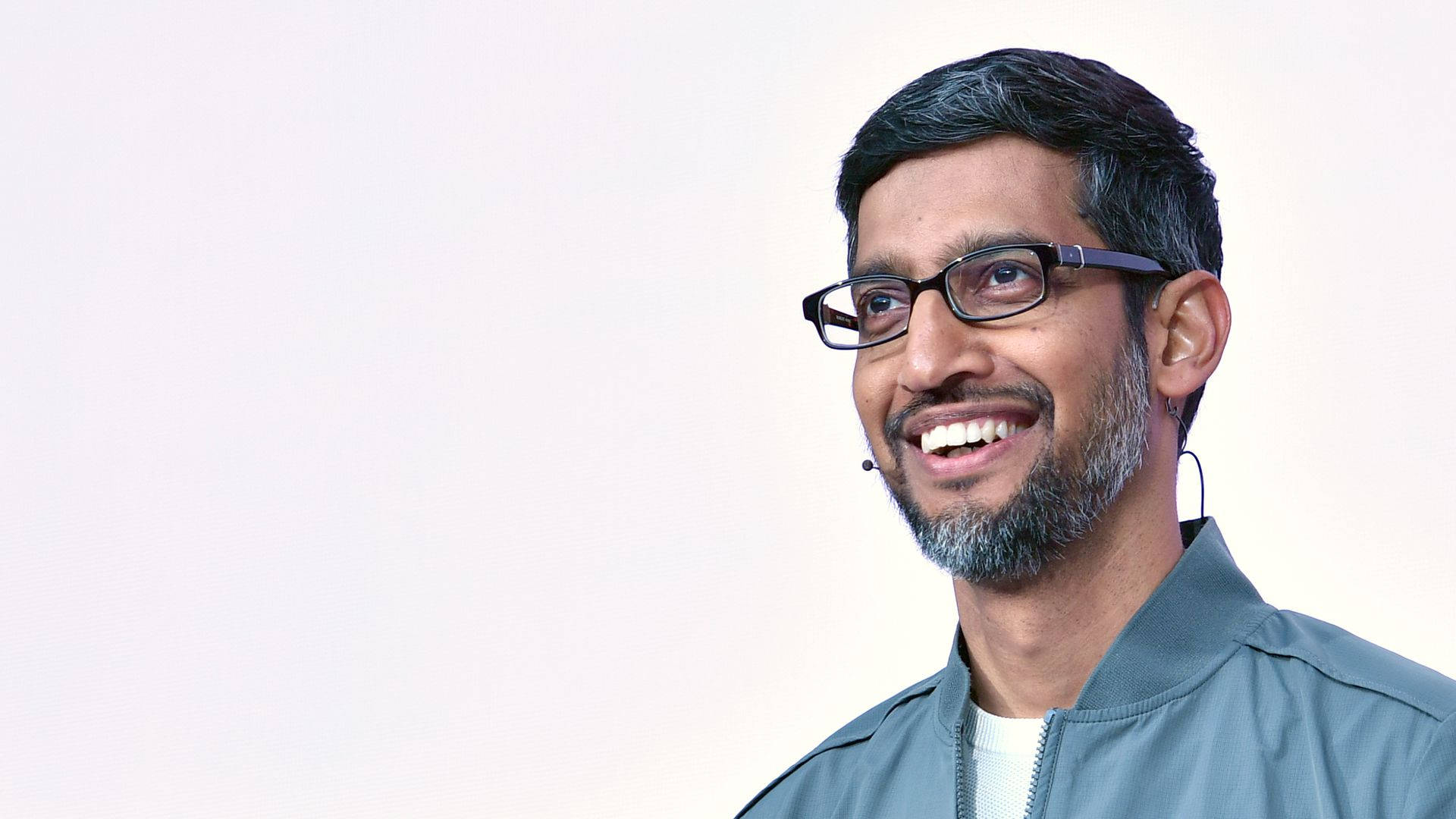 google 桌布 壁紙 — 一位戴眼鏡、留鬍鬚的 smiling 男士，身穿灰色外套，背景為純白色。