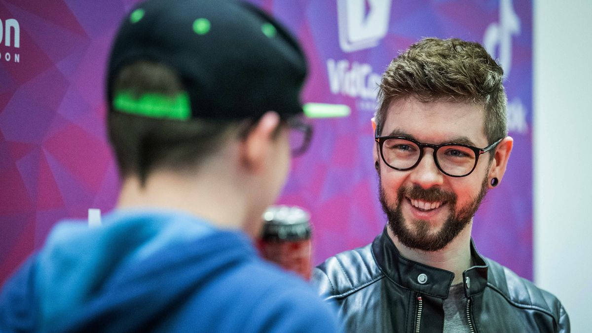 Jacksepticeye 桌布 壁紙 — 一位戴眼鏡、留鬍鬚的男士在充滿活力的紫色品牌活動背景前與另一人交談。