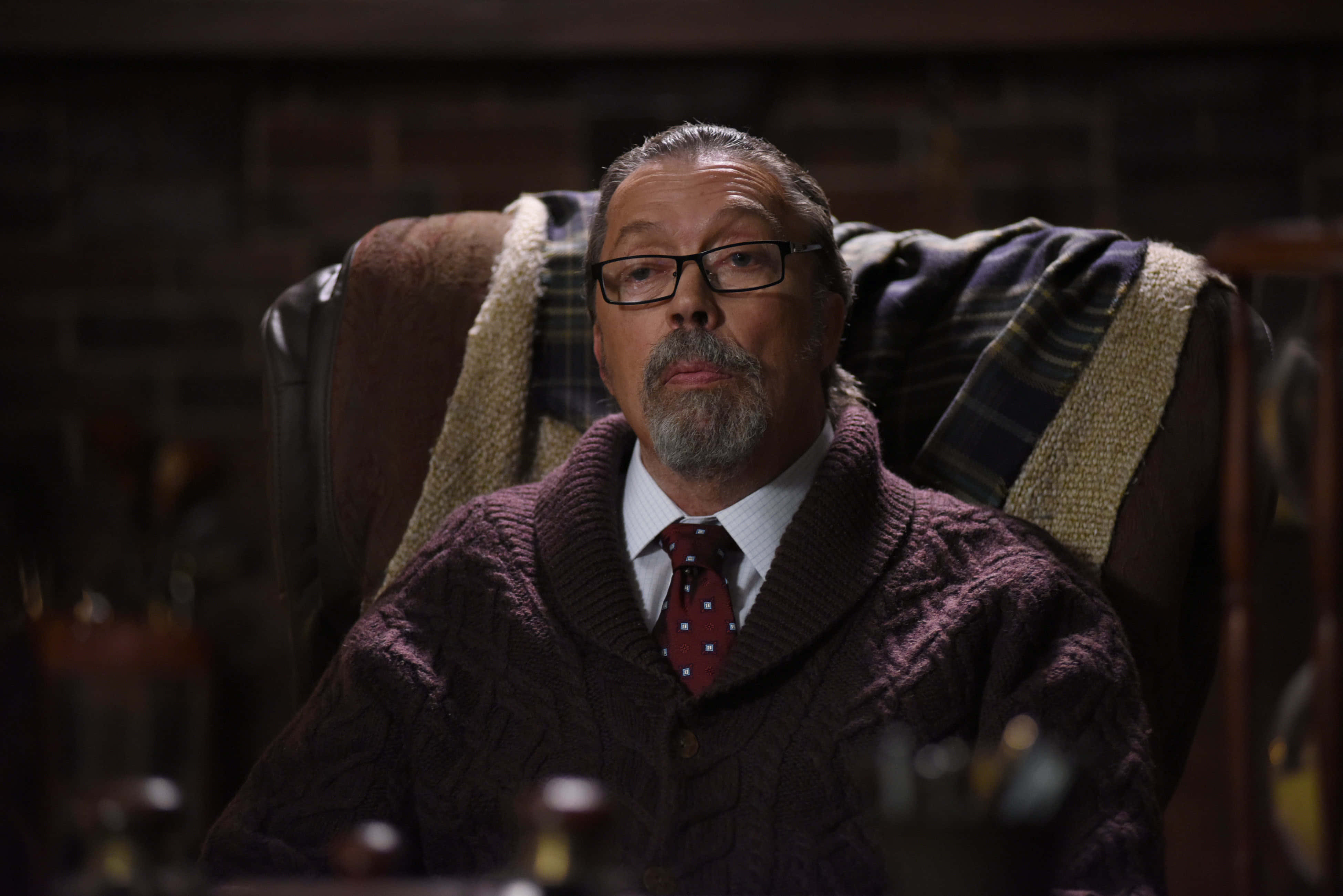 Tim Curry 桌布 壁紙 — 一位戴眼鏡、留鬍鬚的男子坐在皮椅上，身穿棗紅色絞花針織毛衣和紅色領帶，背景是磚牆。