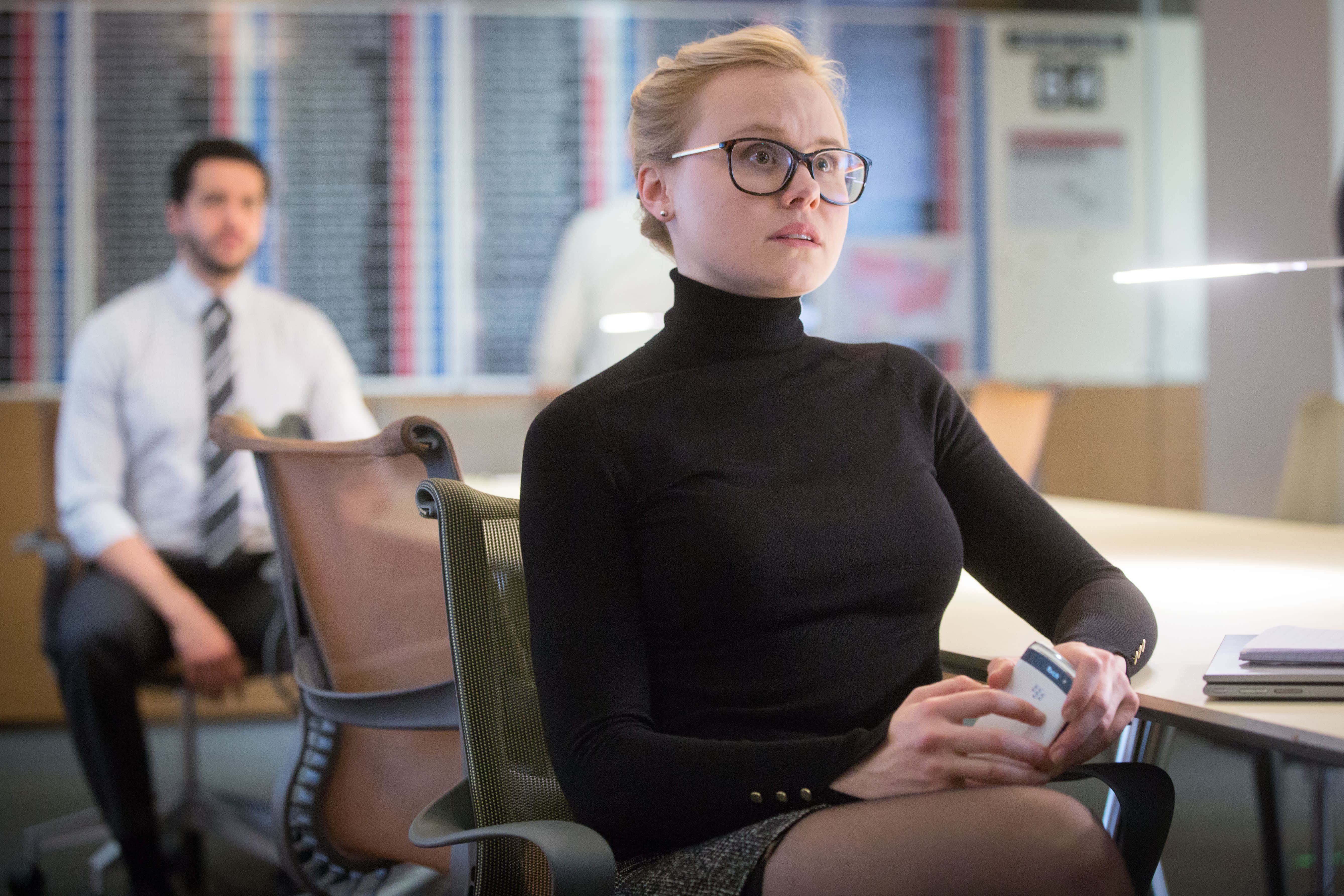 Alison Pill 桌布 壁紙 — 一位戴眼鏡、穿黑色高領毛衣的專注女性坐在現代辦公室的辦公桌前，手持電話，身後一位打領帶的男性模糊地坐著。