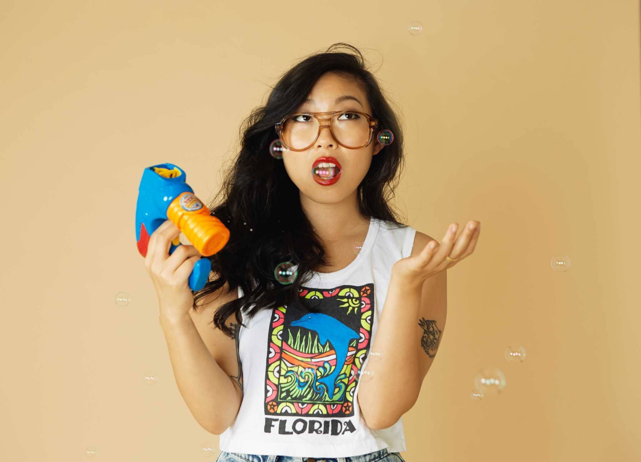 Awkwafina 桌布 壁紙 — 一位戴眼鏡、身穿佛羅里達背心的年輕女性，在米色背景前俏皮地吹泡泡。
