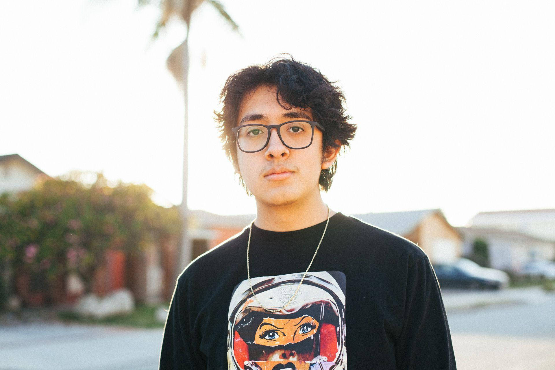 Eyeglasses 桌布 壁紙 — 一位戴眼鏡、身穿圖案T恤的年輕男子站在陽光明媚的郊區街道上，背景中的棕櫚樹和房屋呈現柔和的焦點。
