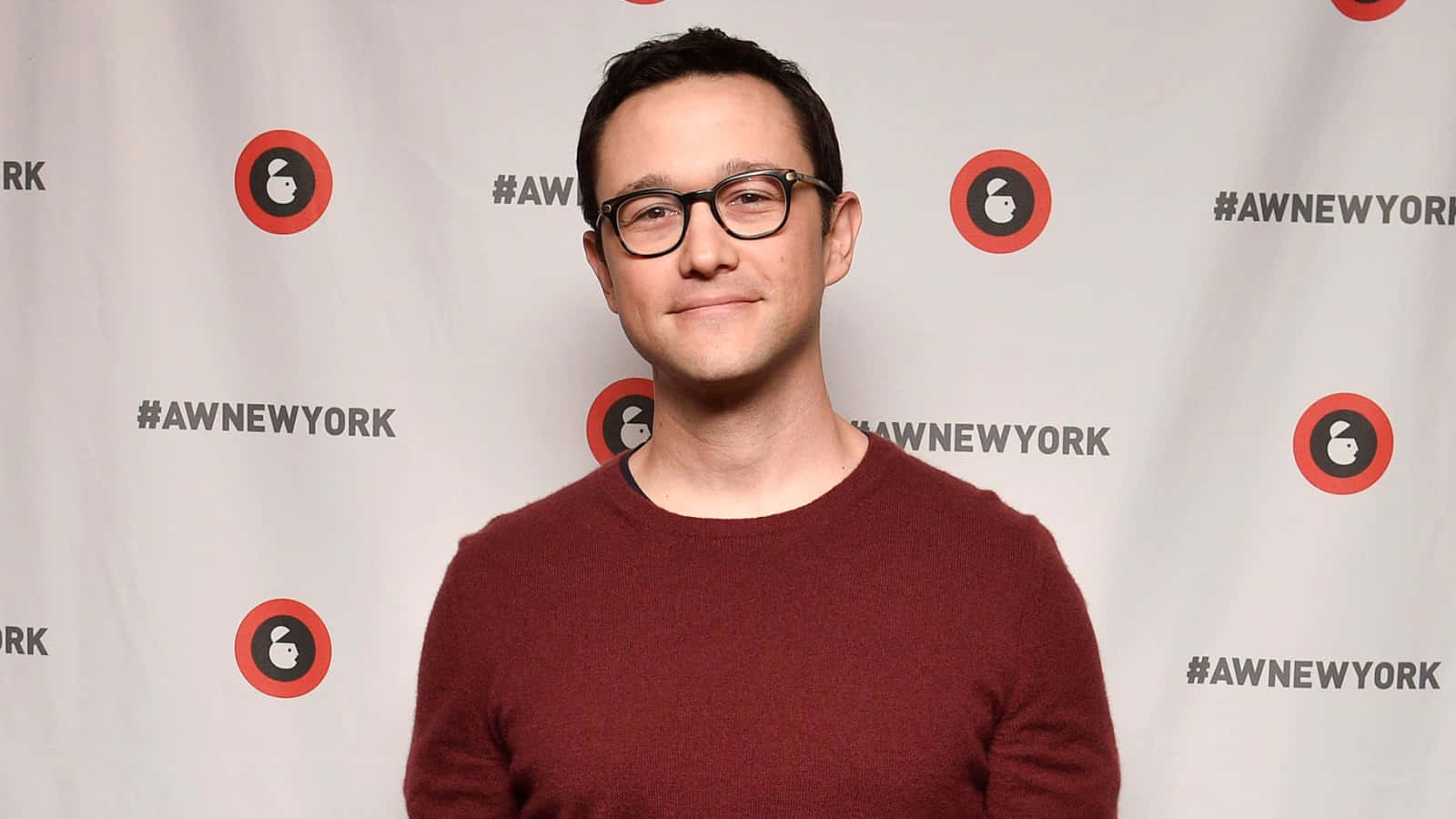 joseph gordon-levitt 桌布 壁紙 — 一位戴眼鏡、身穿栗色毛衣的男子在品牌活動背景前對著鏡頭微笑。