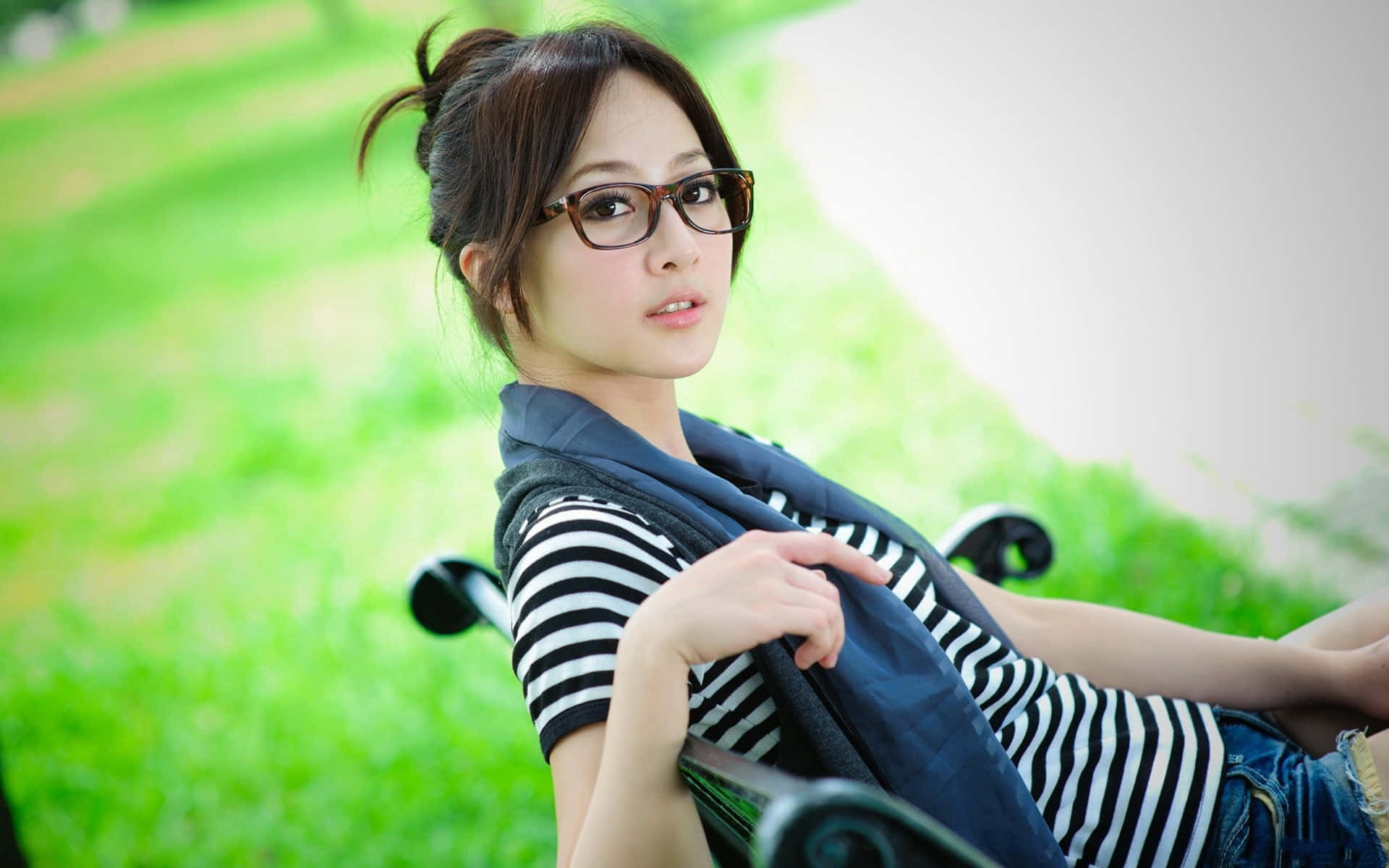 桌布 壁紙 — 一位戴眼鏡、身穿條紋襯衫的年輕女性，在充滿活力的綠色草地背景襯托下，悠閒地躺在公園長椅上。