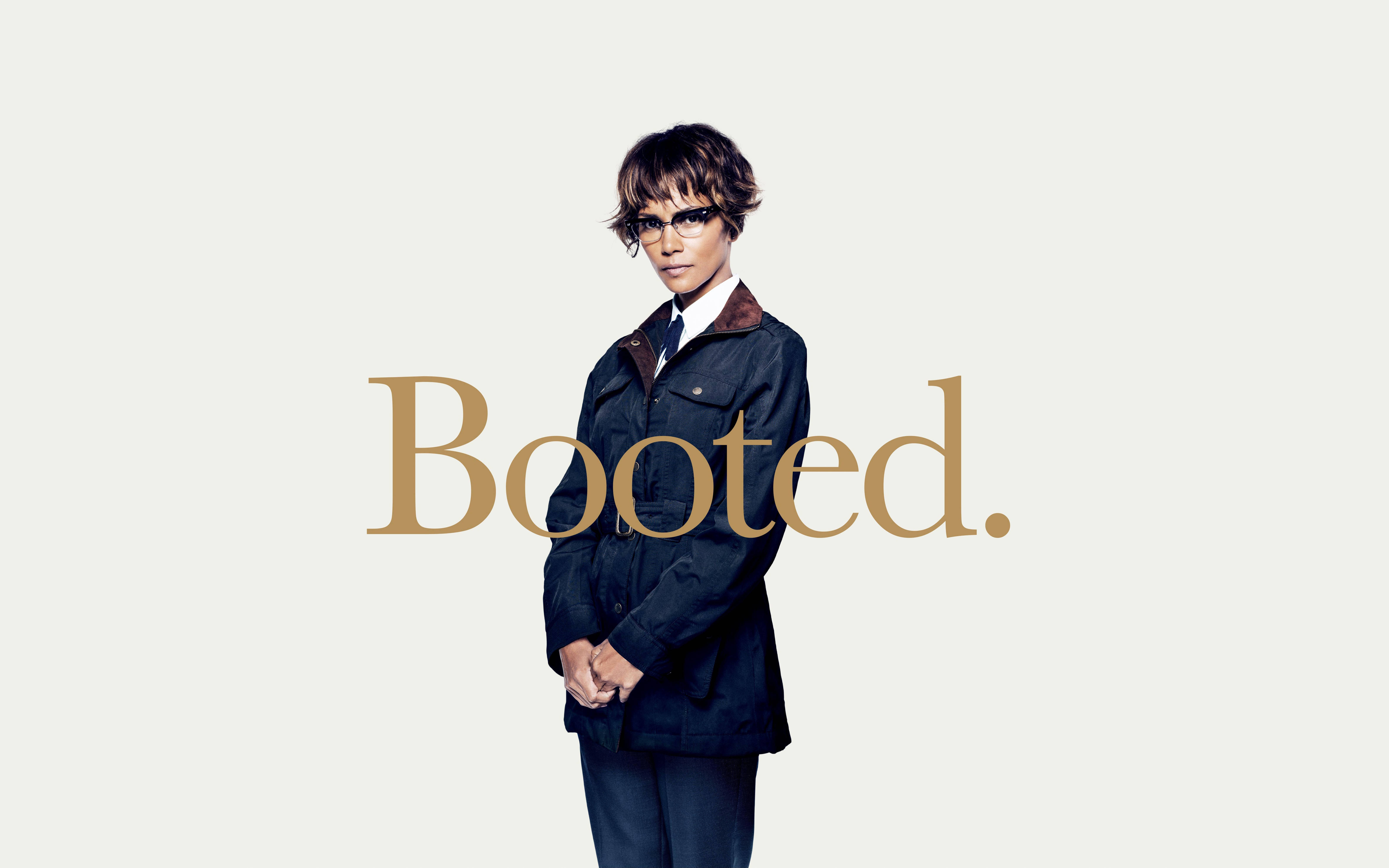 Kingsman 桌布 壁紙 — 一位戴眼鏡、身穿深色外套的年輕人自信地站在素色背景前，背景上有金色文字「Booted.」。