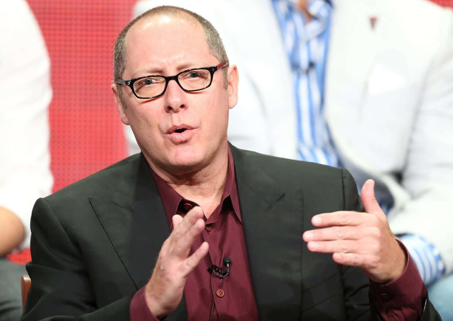 james spader 桌布 壁紙 — 一位戴眼鏡、身穿深色西裝的男子在紅色背景的座談會上發言時做手勢。
