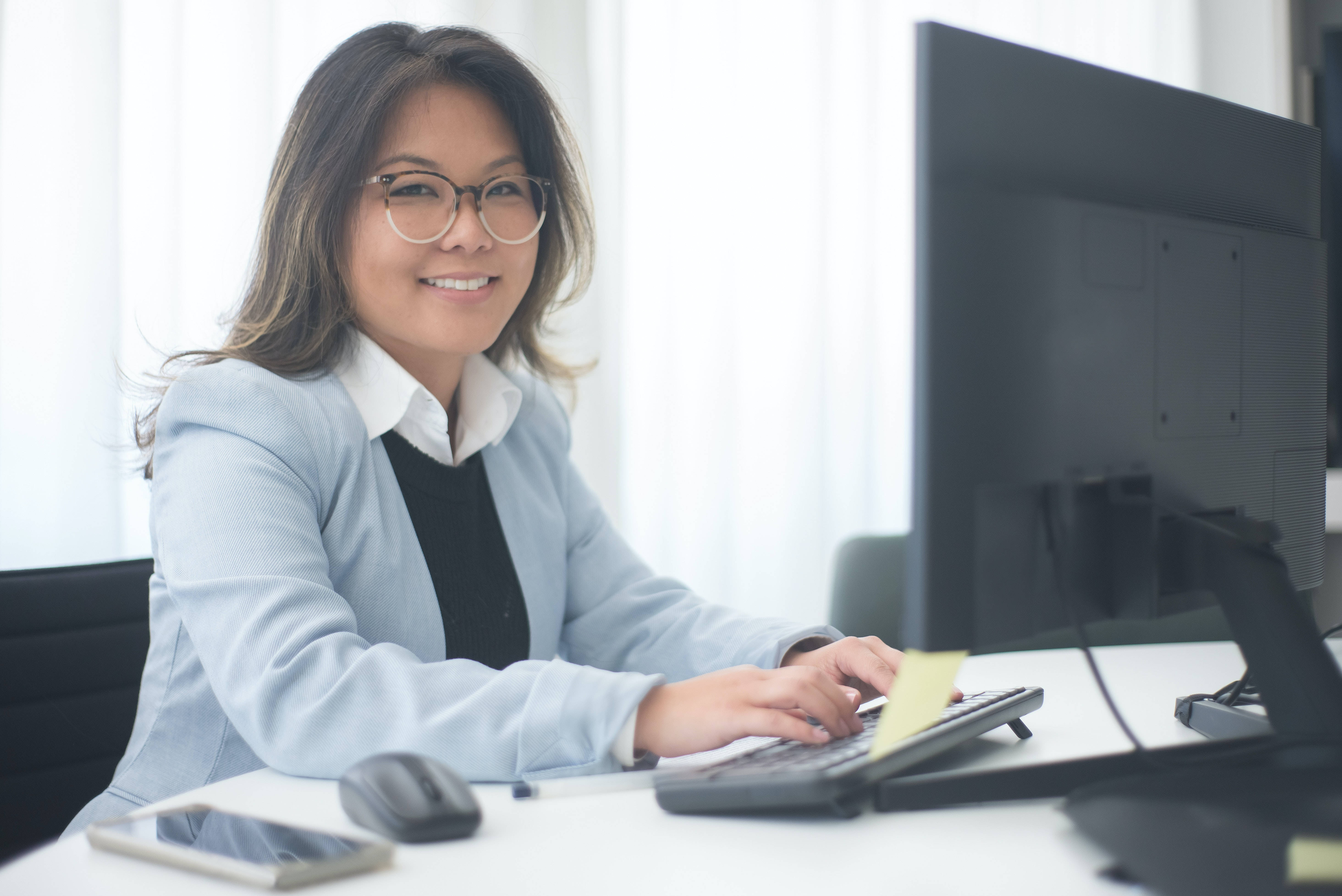 job 桌布 壁紙 — 一位戴眼鏡、身穿淺藍色西裝外套的微笑女性正在辦公桌前打字，附近有電腦顯示器和智慧型手機。