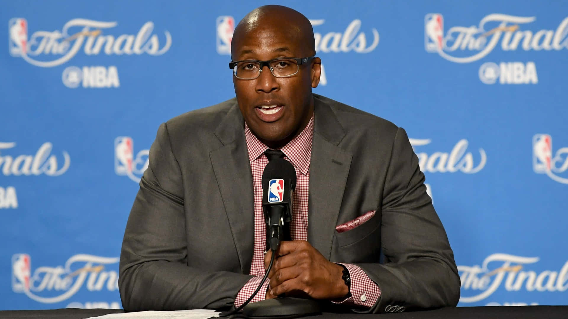 金州勇士 桌布 壁紙 — 一位戴眼鏡、身穿灰色西裝的光頭黑人男子在NBA總決賽記者會上對著麥克風發言。
