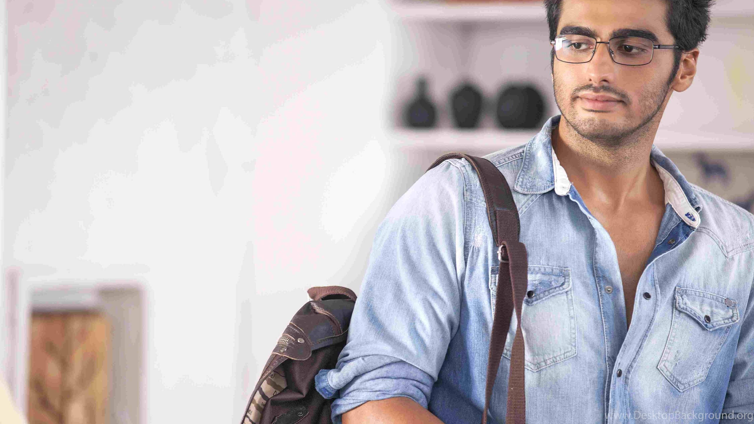 Arjun Kapoor 桌布 壁紙 — 一位戴眼鏡、身穿牛仔襯衫的年輕男子，在柔和光線的室內環境中背著棕色背包，視線望向一旁。