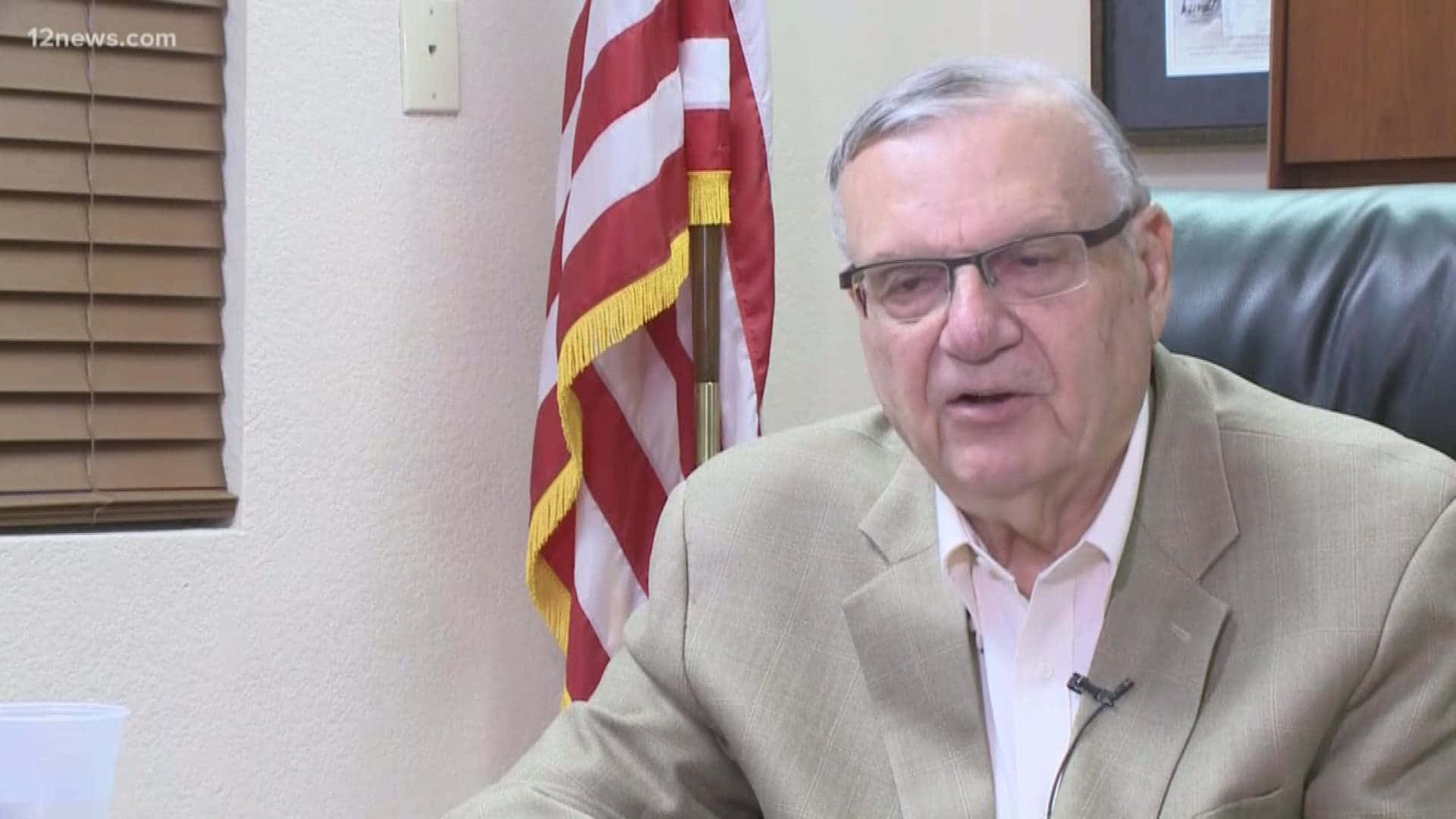 政治 桌布 壁紙 — 一位戴眼鏡、身穿米色西裝的年長男性坐在辦公室的皮椅上，身後有一面美國國旗。