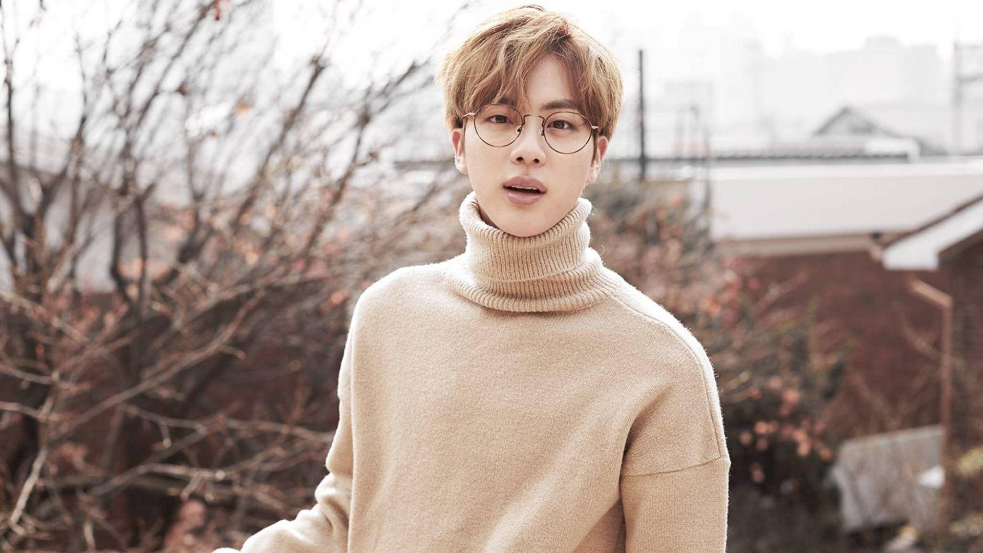 kim seok jin 桌布 壁紙 — 一位戴眼鏡、身穿米色高領毛衣的年輕男子站在戶外，背景是光禿的樹枝和柔和冬日光線。