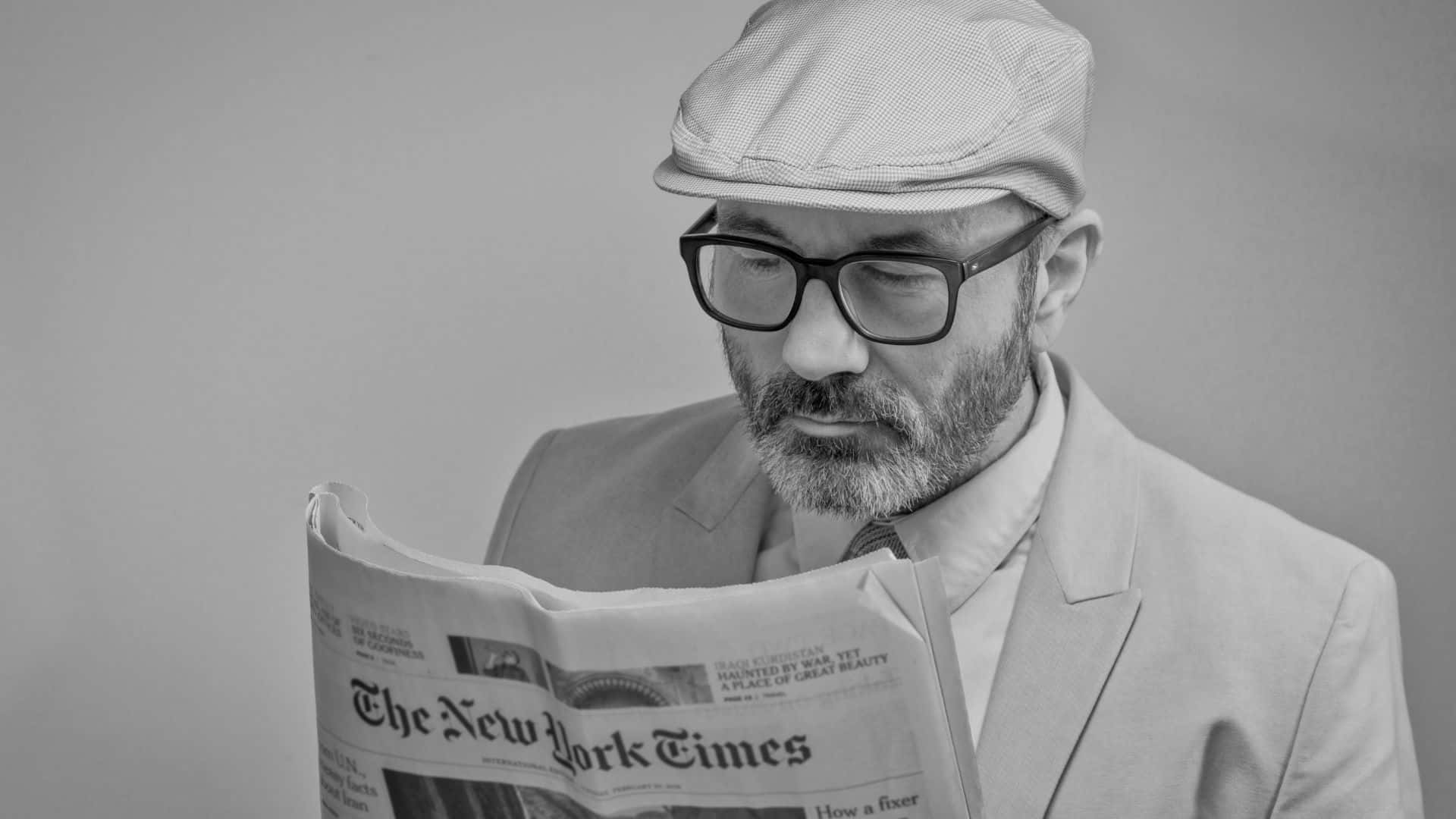 Black And White Newspaper 桌布 壁紙 — 一位戴眼鏡、身穿西裝和帽子的男士，在純灰色背景前以單色閱讀《紐約時報》。