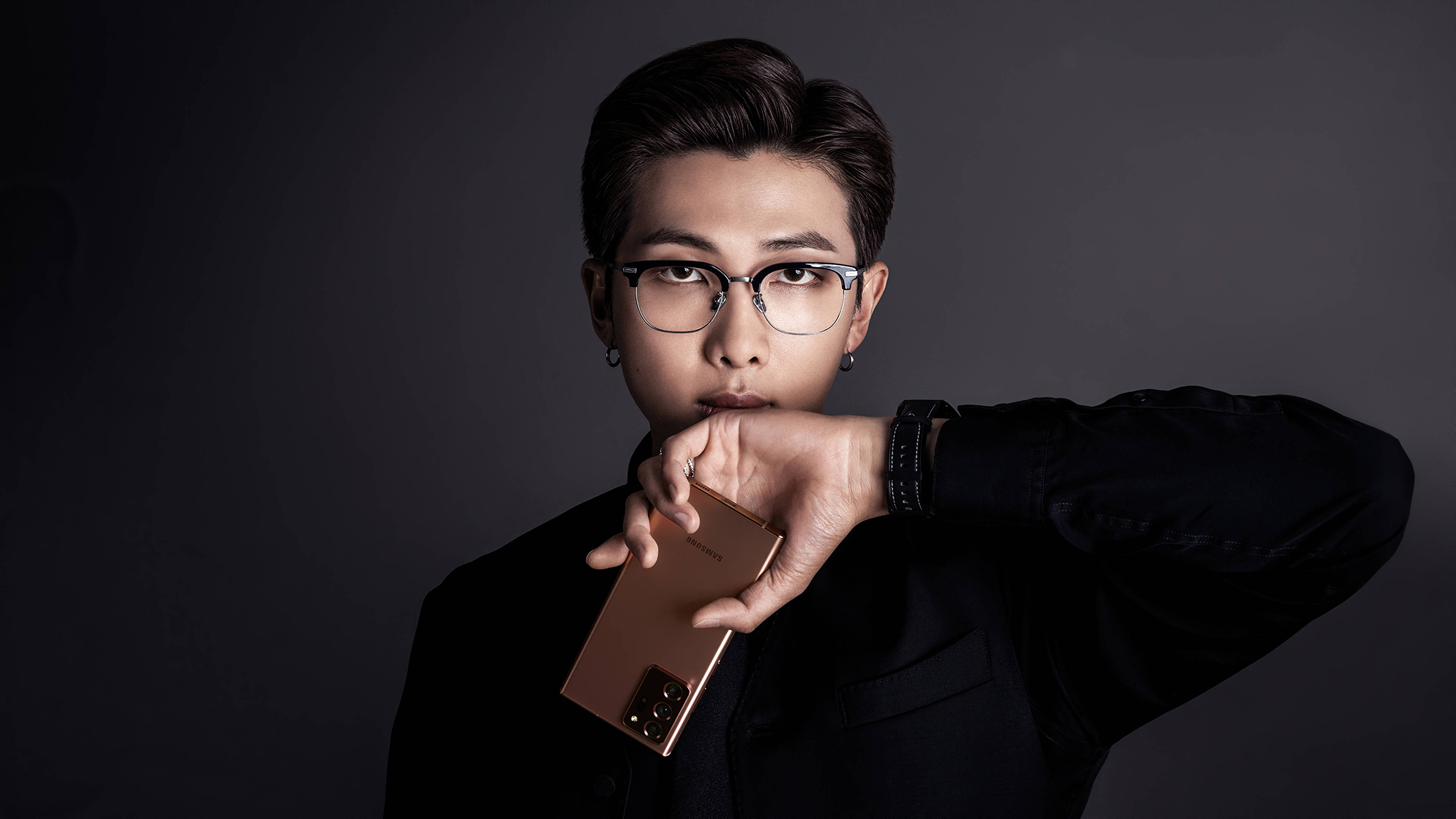 美學 BTS 桌布 壁紙 — 一位戴眼鏡、身穿黑色襯衫的年輕男子，在深灰色工作室背景前手持玫瑰金色智慧型手機。