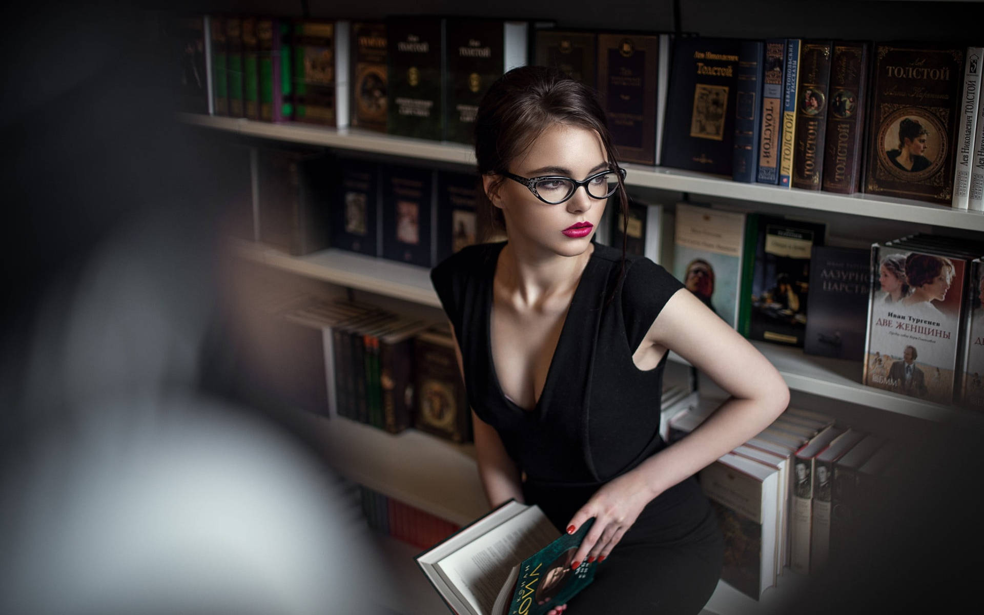 Reading 桌布 壁紙 — 一位戴眼鏡、身穿黑色連衣裙的女性在光線昏暗的圖書館裡拿著一本書，身後是書架。