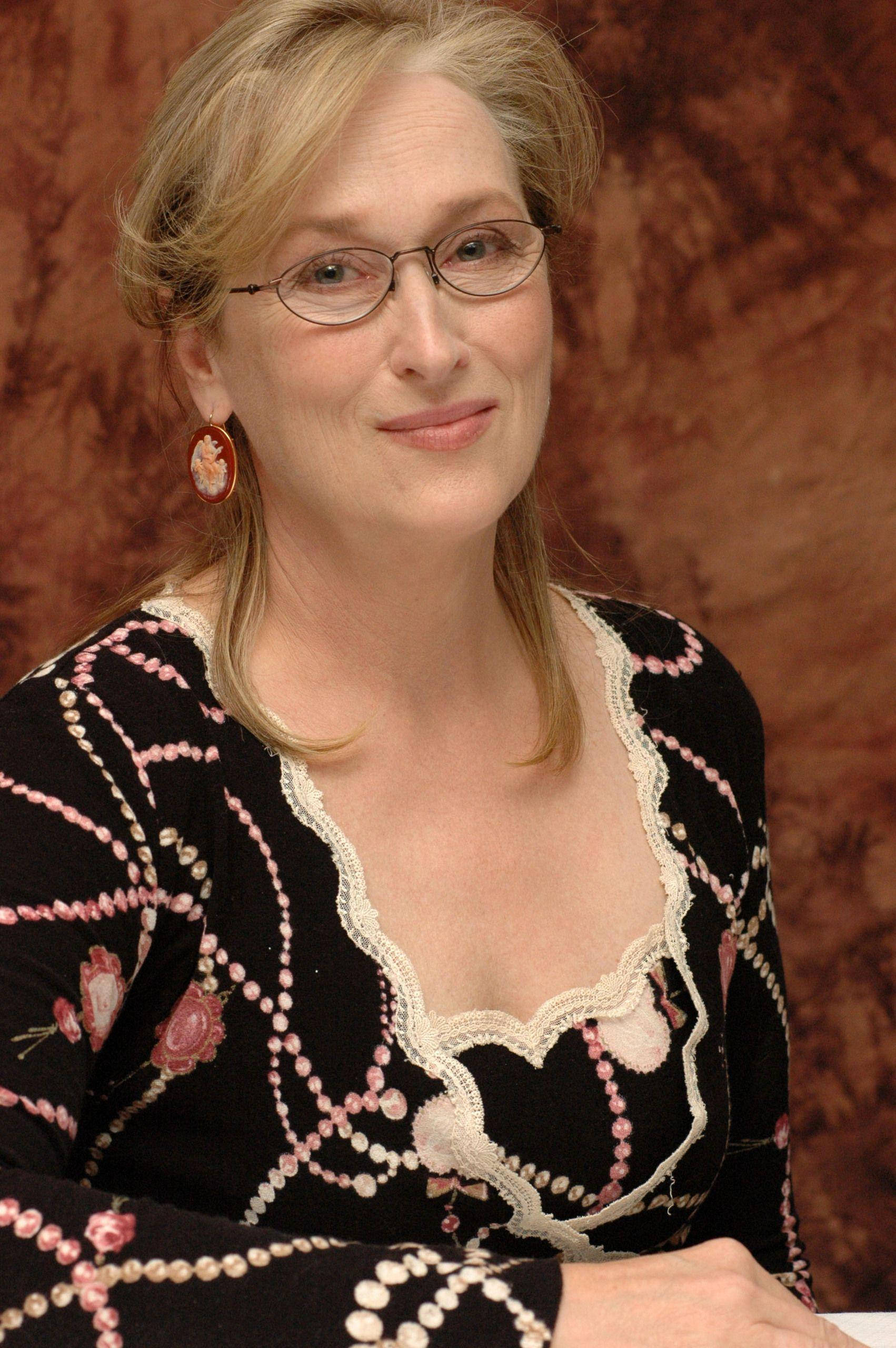 Meryl Streep 桌布 壁紙 — 一位戴眼鏡、金髮的女性，穿著帶有粉紅色花卉刺繡的黑色圖案上衣，在質感棕色背景前溫柔微笑。