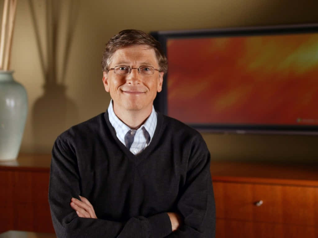 Bill Gates 桌布 壁紙 — 一位戴眼鏡、雙臂交叉的微笑男子自信地站在一個溫暖照明的室內環境中，身後有木製家具和抽象藝術品。