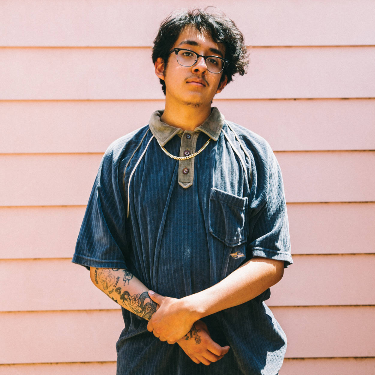 Cuco 桌布 壁紙 — 一位戴眼鏡及有刺青的年輕男子自信地站在柔軟的粉紅色木牆前，身穿藍色條紋襯衫。