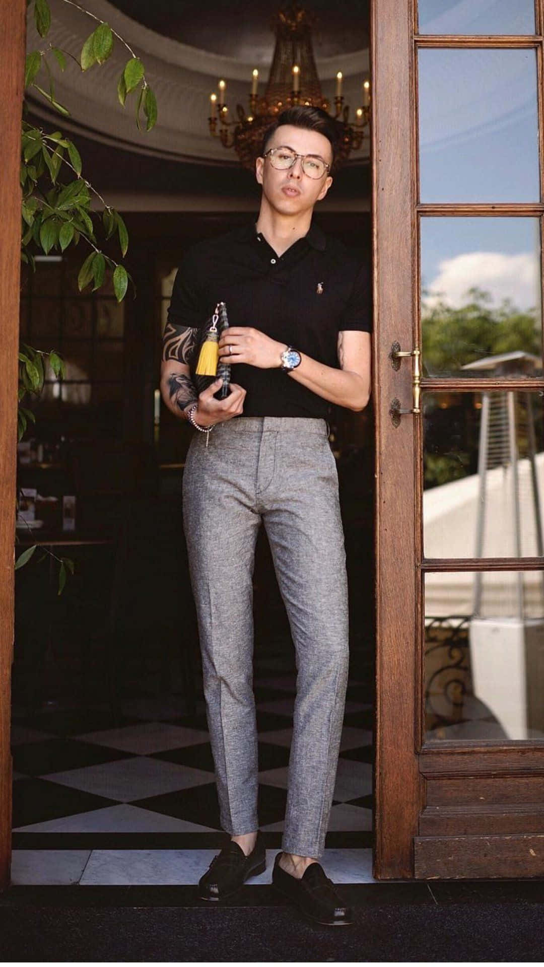 Black Clothing 桌布 壁紙 — 一位戴眼鏡及有刺青的時尚男士站在門口，手持酒瓶，背景為木門及吊燈。