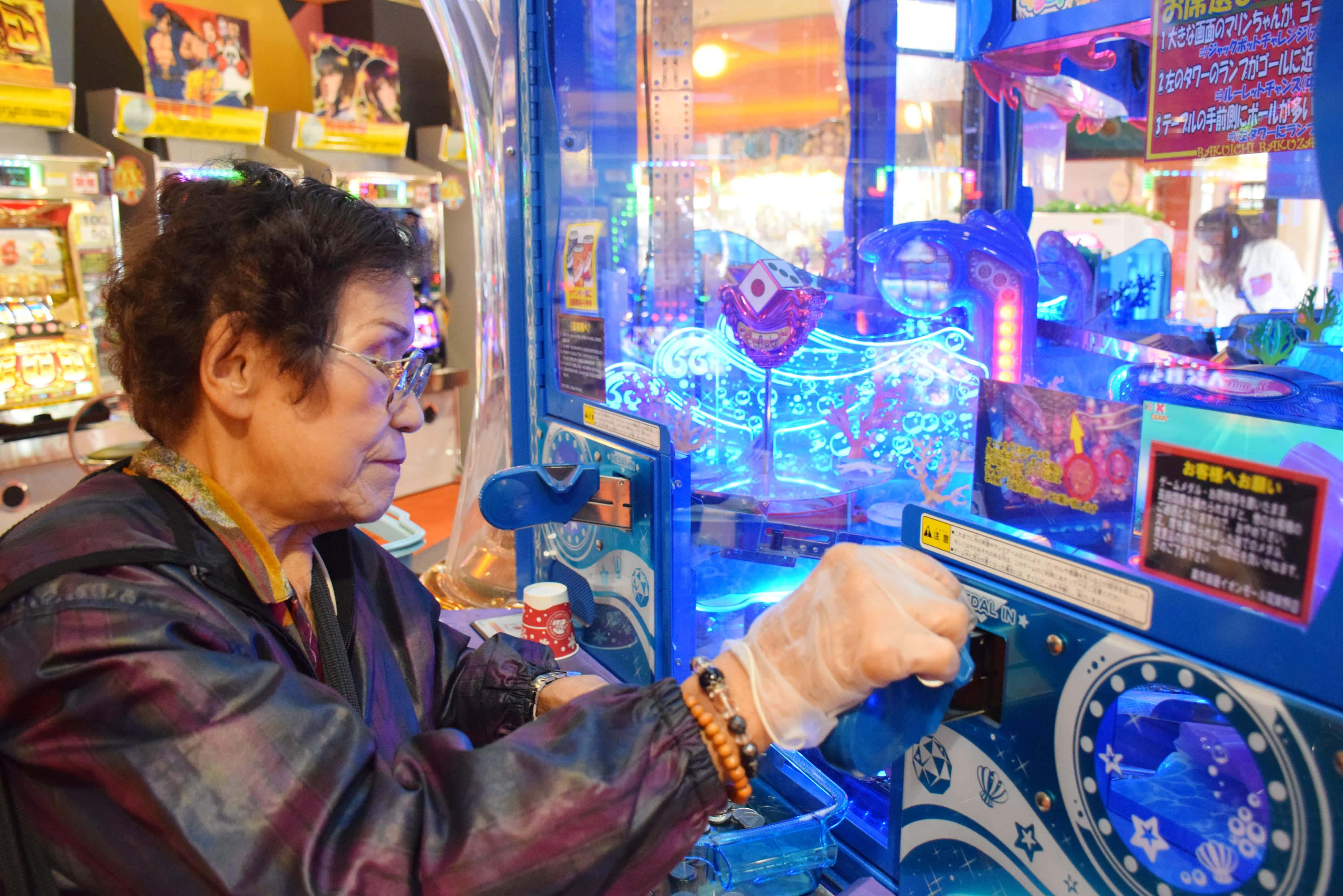 Arcade Games 桌布 壁紙 — 一位戴眼鏡和塑膠手套的老年女士，在熱鬧的遊戲機中心專注地玩著發光的藍色夾娃娃機。