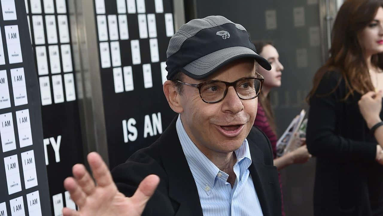 rick moranis 桌布 壁紙 — 一位戴眼鏡和帽子的微笑男子，在重複白色文字的黑色牆前熱情地做手勢。