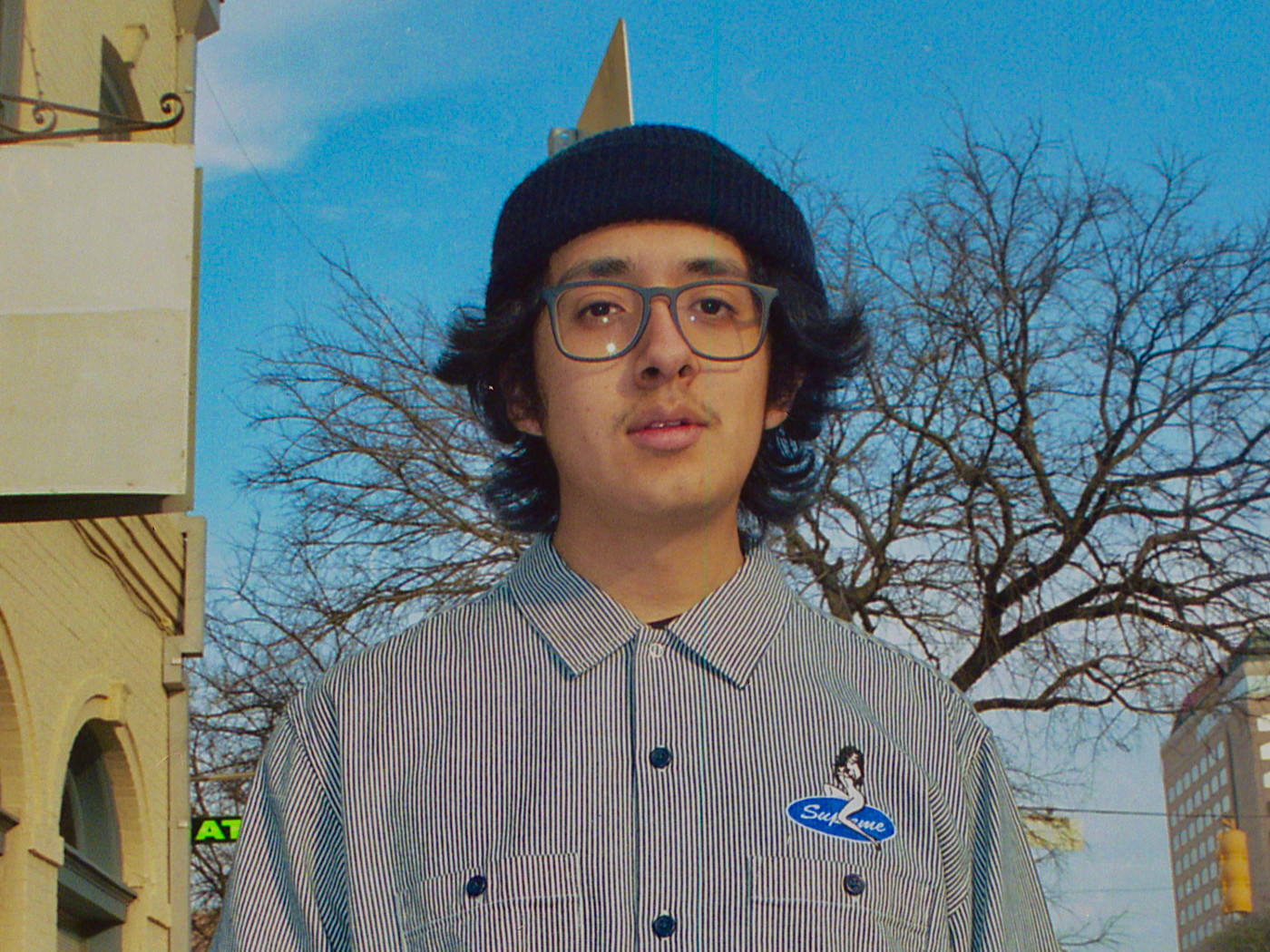 Cuco 桌布 壁紙 — 一位戴眼鏡和毛帽的年輕男子站在戶外，背景是晴朗的藍天、光禿的樹木和城市建築。