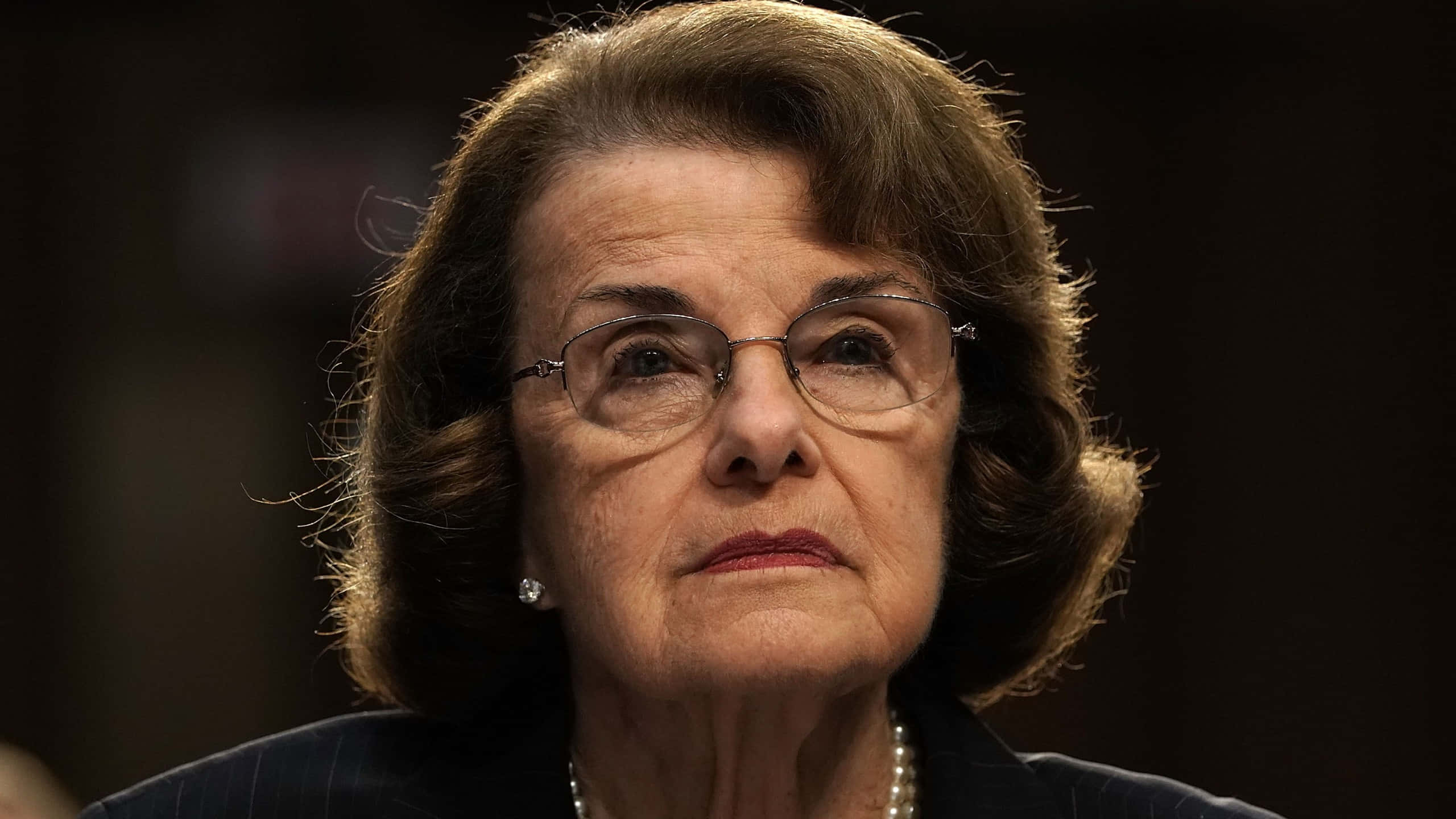 Dianne Feinstein 桌布 壁紙 — 一位戴眼鏡和珍珠項鍊的老年婦女，表情嚴肅地向上看的特寫肖像。