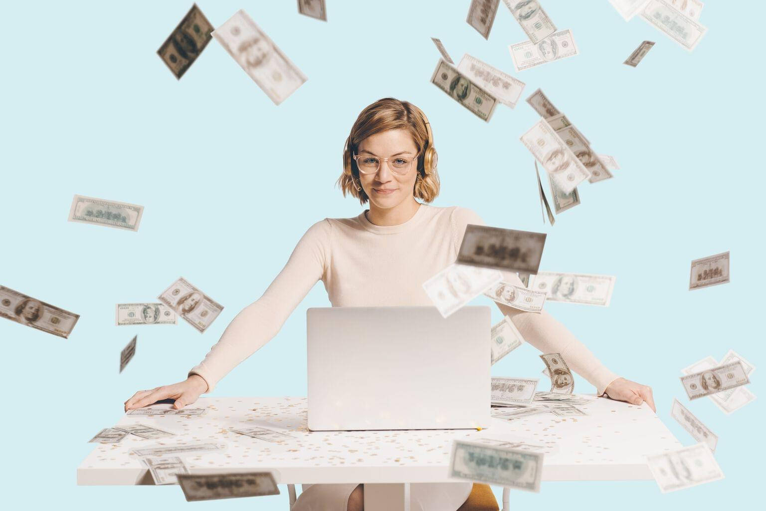金錢 桌布 壁紙 — 一位戴眼鏡和耳機的女性微笑著，美元鈔票如雨般落在她周圍的白色桌子上，桌上放著一部筆記型電腦。