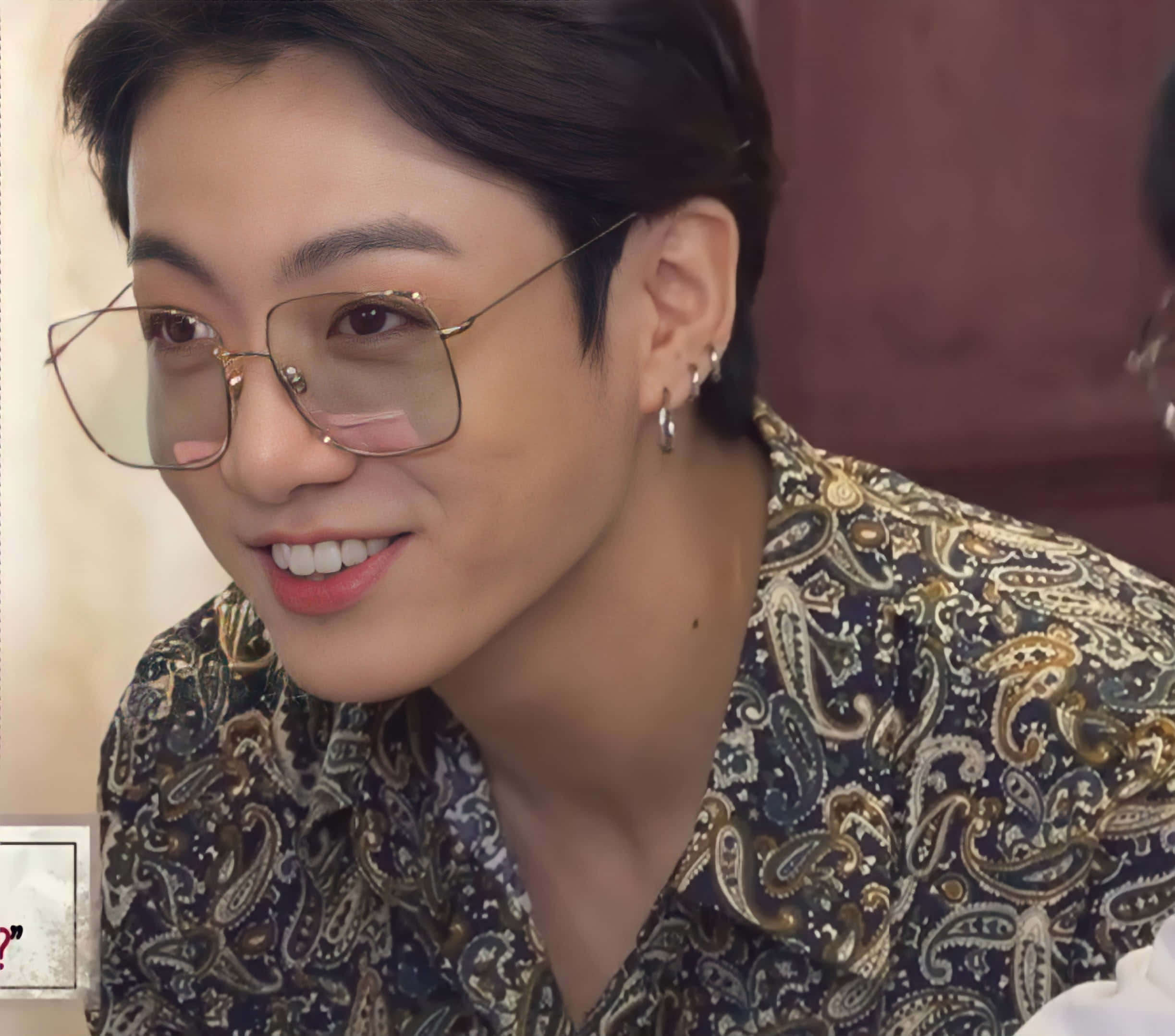Jungkook 桌布 壁紙 — 一位戴眼鏡和耳環的年輕男子穿著佩斯利圖案襯衫，溫暖地微笑。