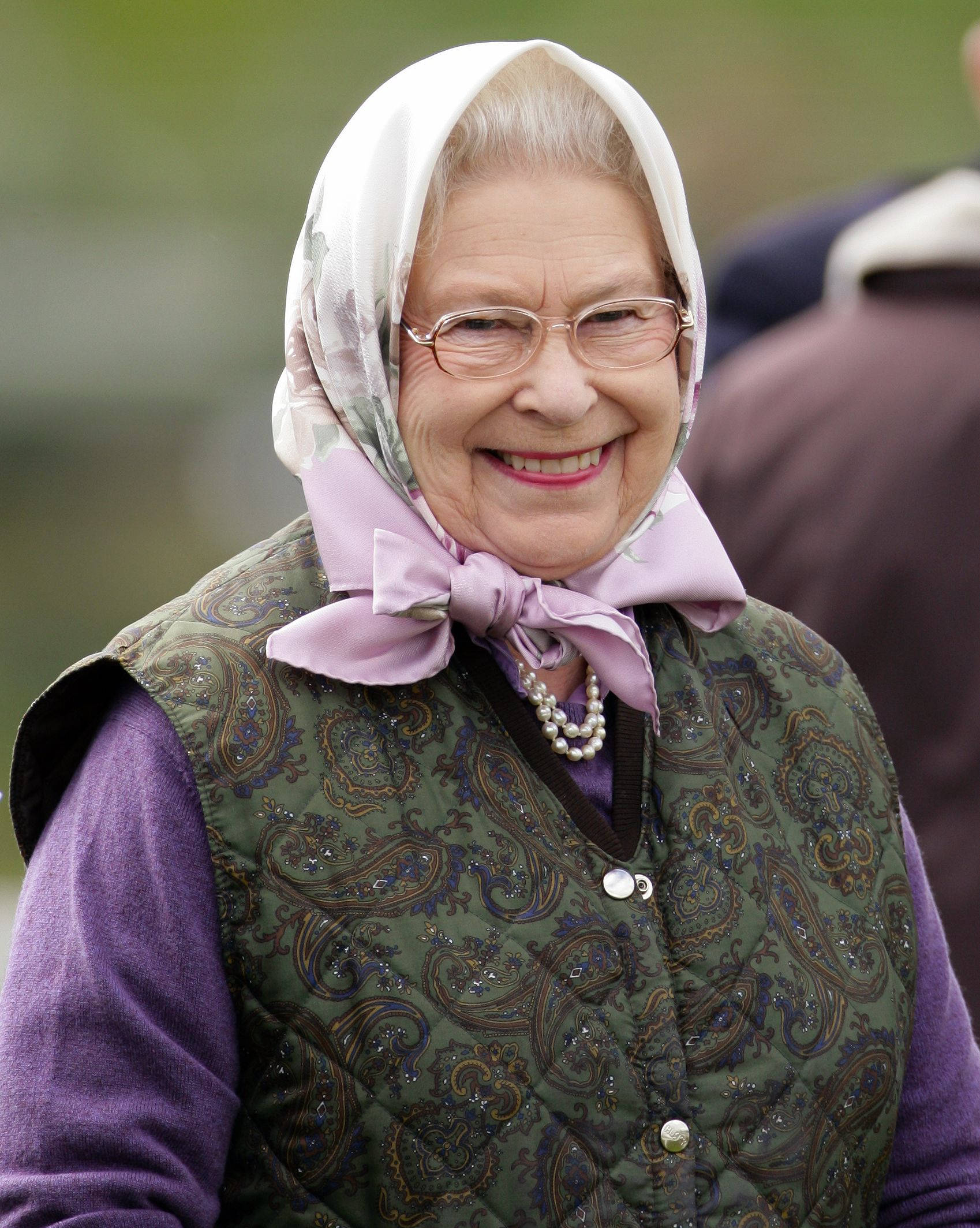 Queen Elizabeth 桌布 壁紙 — 一位戴眼鏡和花卉頭巾的老婦人，穿著圖案背心和珍珠項鍊，溫暖地微笑。