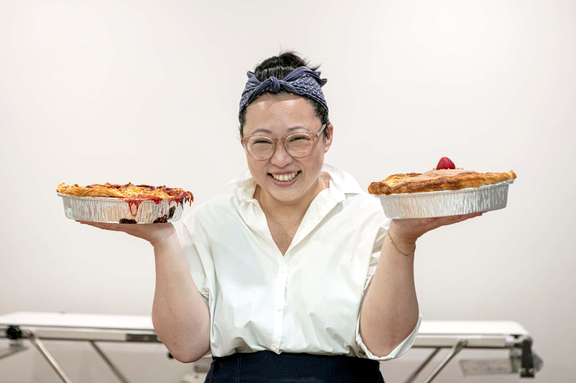 人物 桌布 壁紙 — 一位戴眼鏡和頭巾的開朗女士，自豪地捧著兩個剛出爐的錫箔盤餡餅，笑容燦爛。