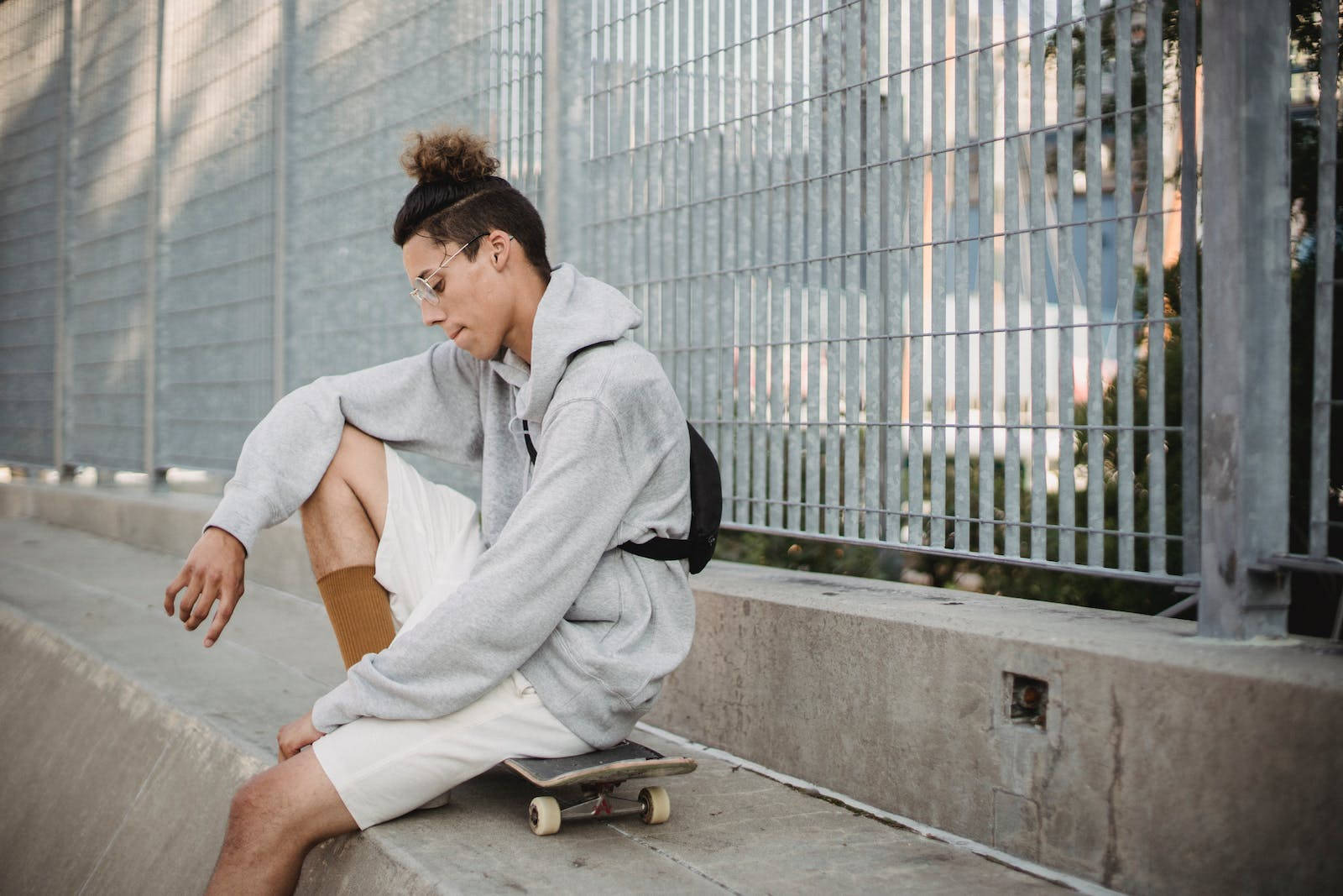 Cool Sad 桌布 壁紙 — 一位戴眼鏡和髮髻的年輕人，在都市日光下靠著金屬圍欄沉思地坐在滑板上。