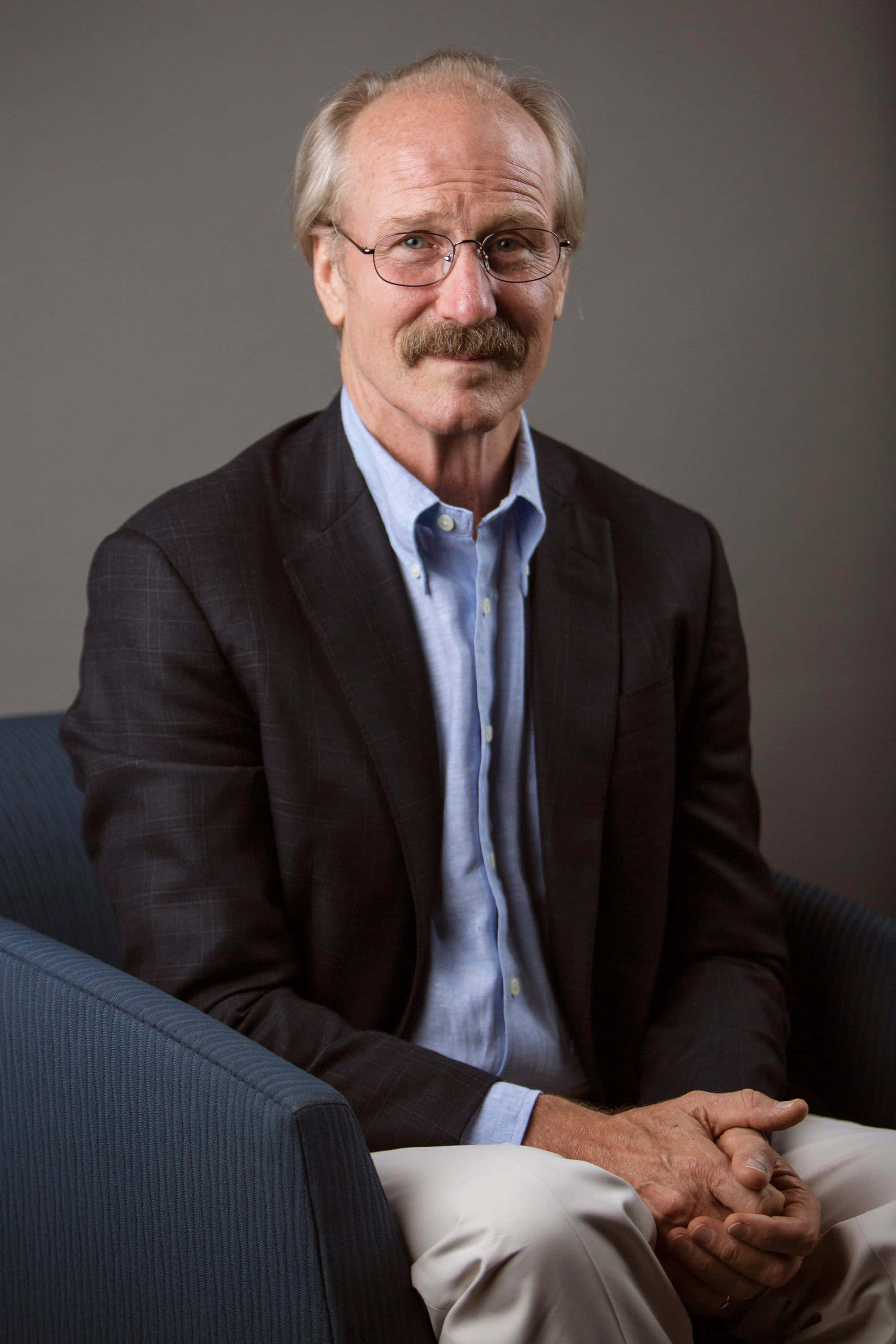William Hurt 桌布 壁紙 — 一位戴眼鏡和鬍子的年長男士，身穿深色西裝外套和淺藍色襯衫，平靜地坐在藍色扶手椅上。