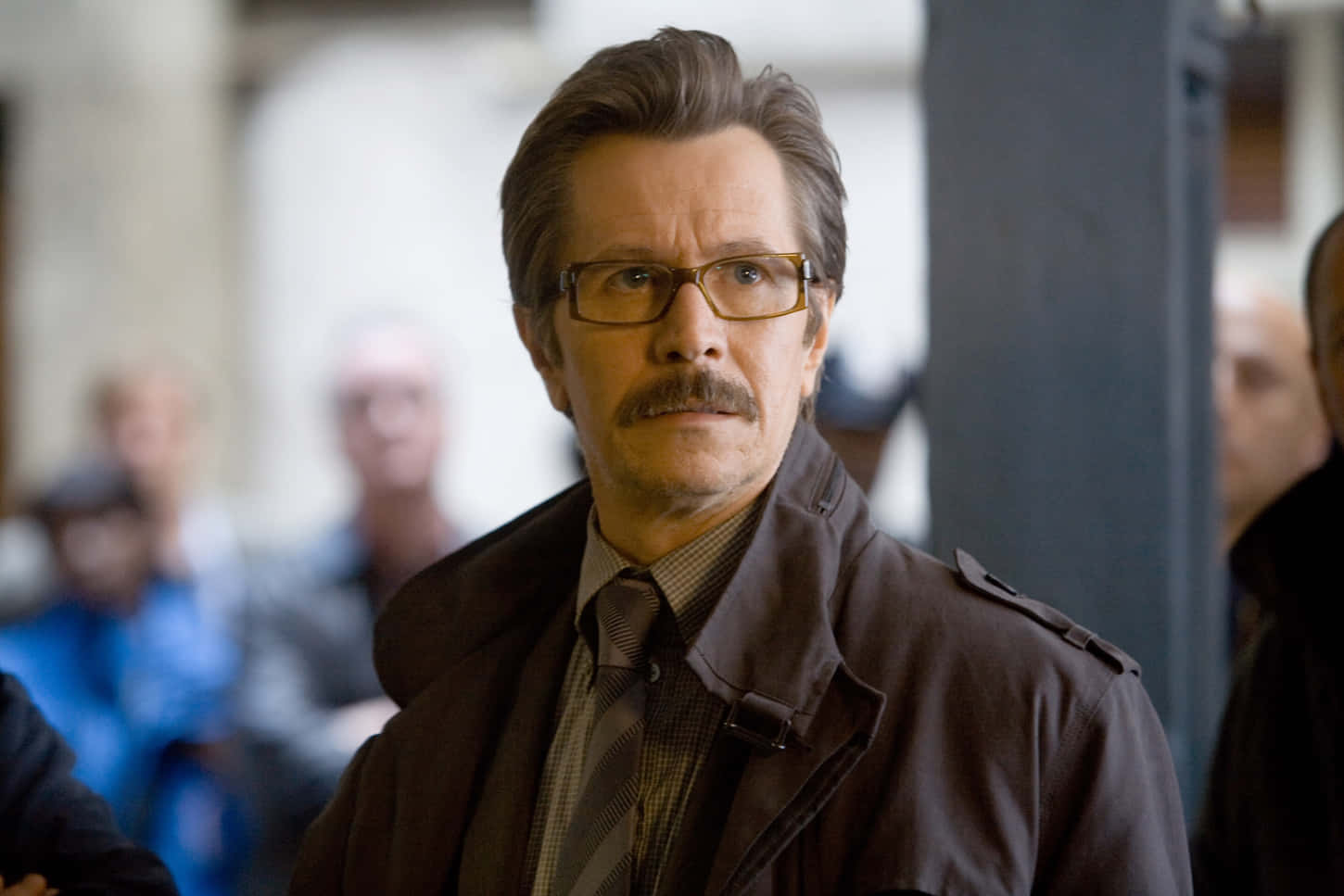 Commissioner Gordon 桌布 壁紙 — 一位戴眼鏡和鬍子的男士穿著棕色風衣和領帶，在模糊的室內環境中專注地向前看。