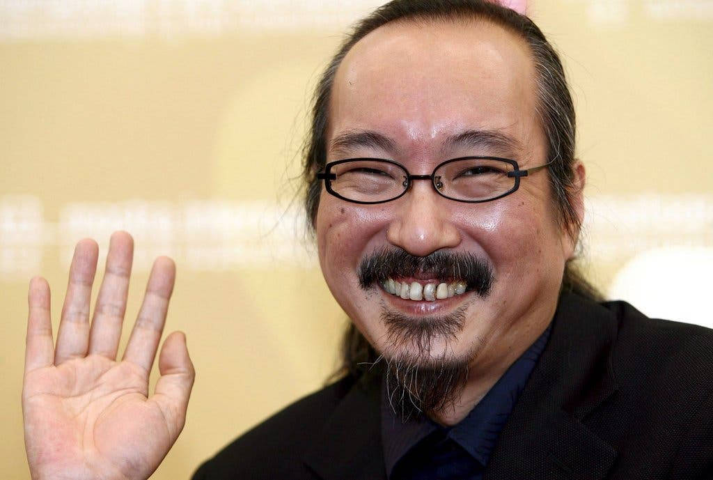 satoshi kon 桌布 壁紙 — 一位戴眼鏡和鬍子的開朗男士，在正式場合中熱情地向鏡頭揮手。