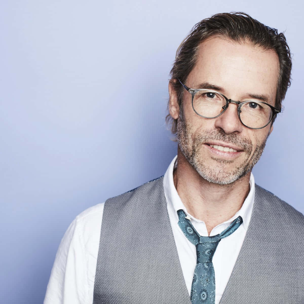 Guy Pearce 桌布 壁紙 — 一位戴眼鏡和鬍鬚的中年男子在柔和的藍色工作室背景前溫暖地微笑。