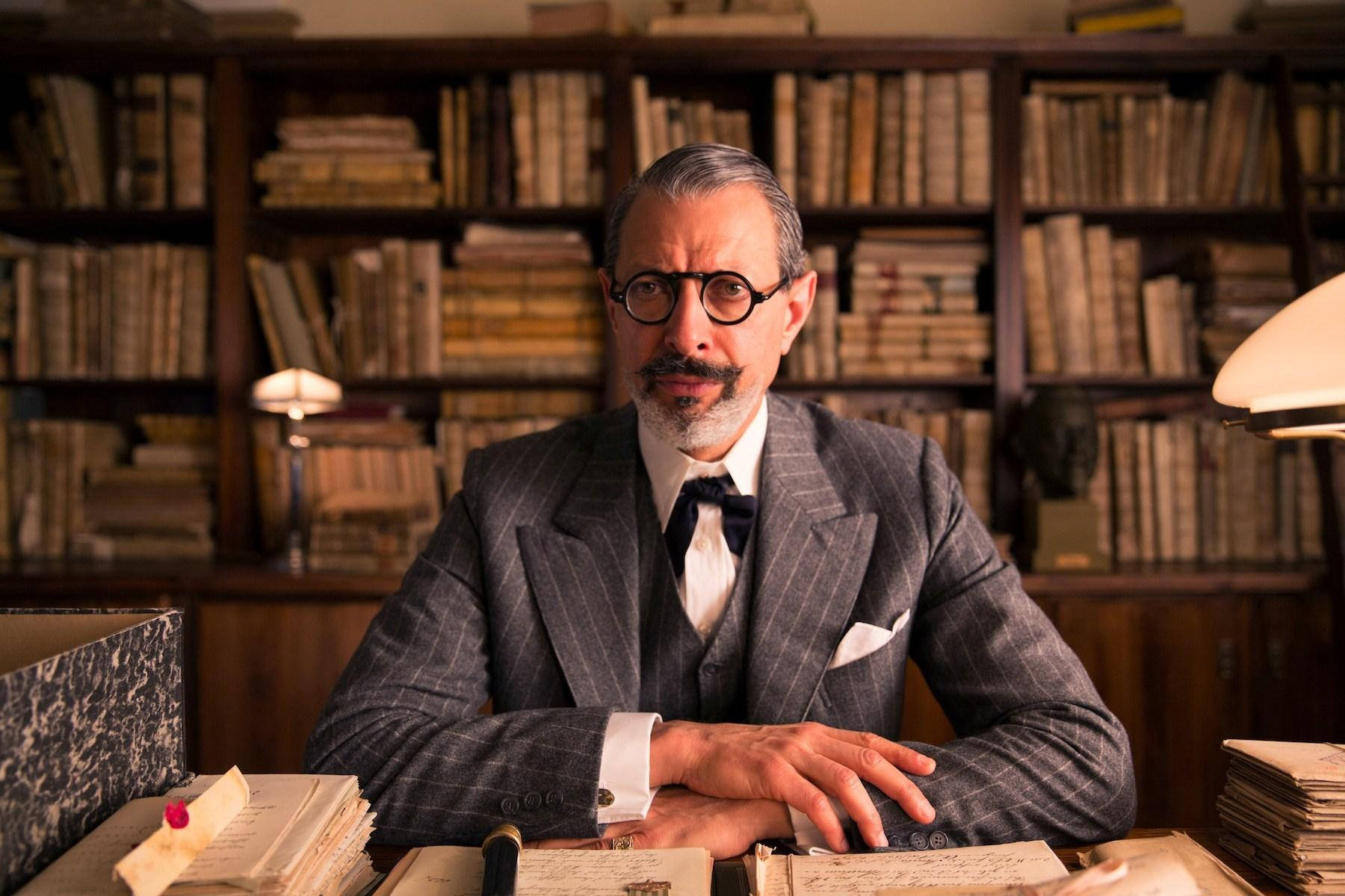 jeff goldblum 桌布 壁紙 — 一位戴眼鏡和鬍鬚的尊貴男士坐在光線昏暗的圖書館中，周圍是高聳的書架，桌上凌亂。