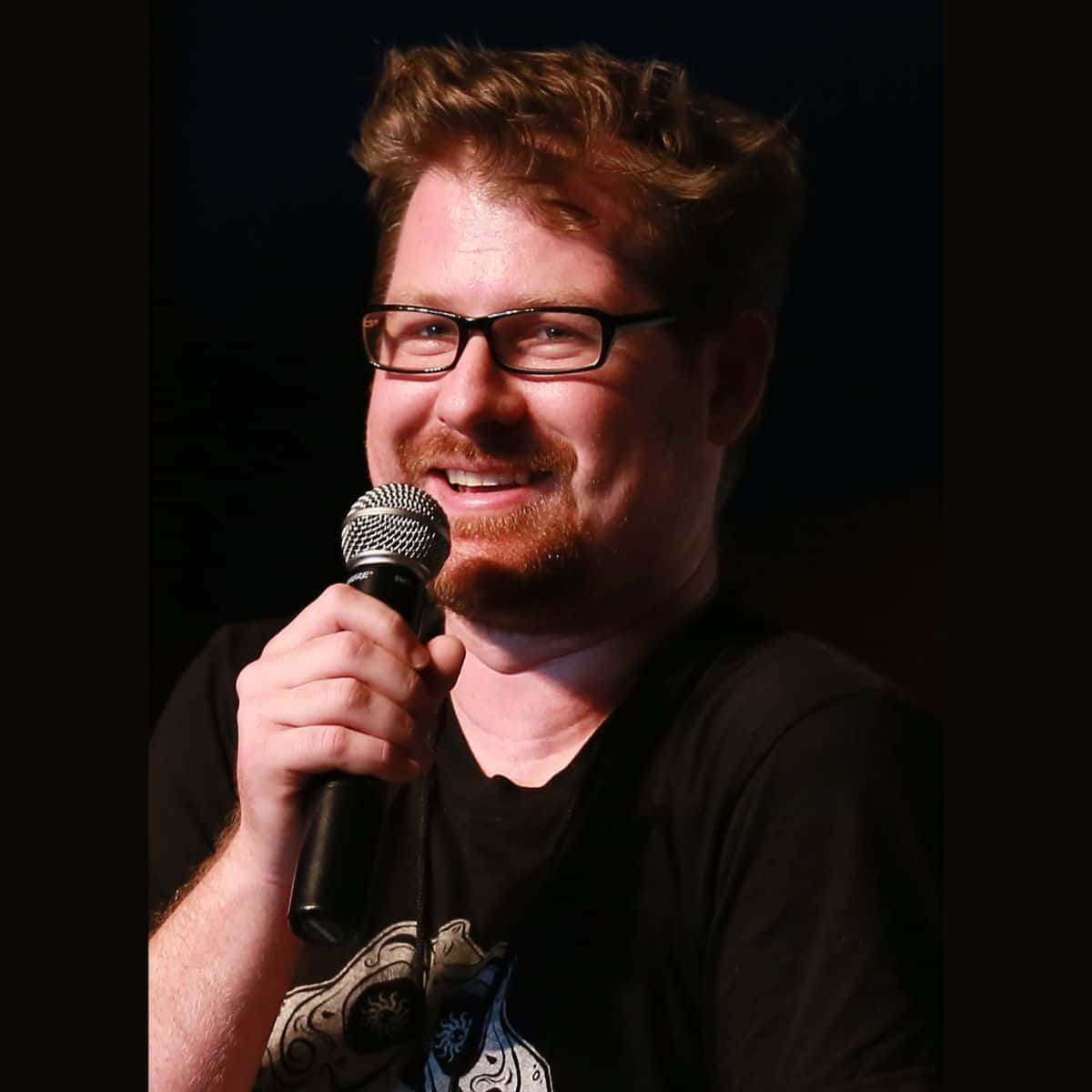 Justin Roiland 桌布 壁紙 — 一位戴眼鏡和鬍鬚的微笑男子在深色背景前拿著麥克風。