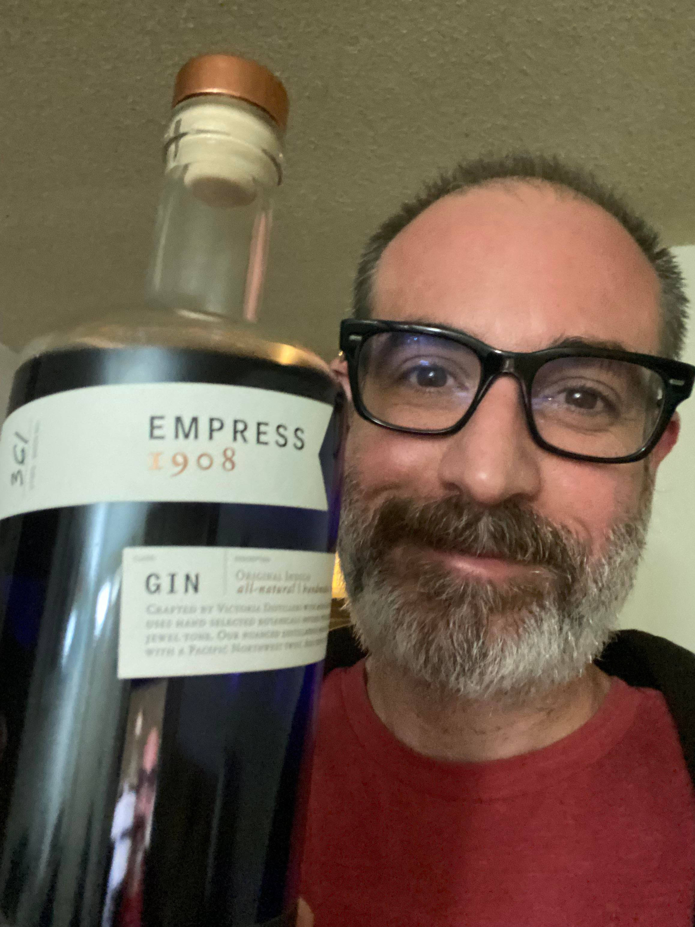 selfie 桌布 壁紙 — 一位戴眼鏡和鬍鬚的男人微笑著，在室內拿著一大瓶Empress 1908琴酒。