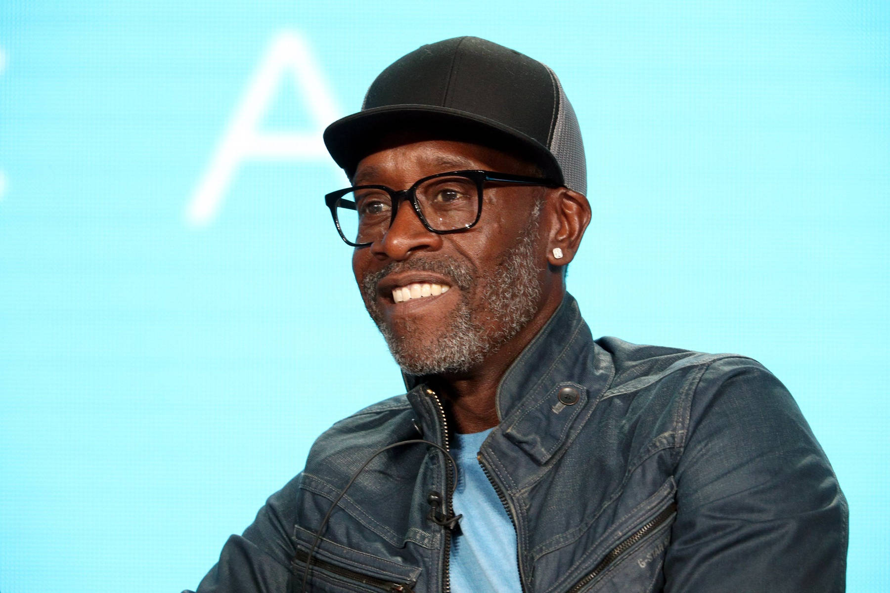 Don Cheadle 桌布 壁紙 — 一位戴眼鏡和黑色帽子的微笑男士，穿著牛仔外套，背景是明亮的藍色。