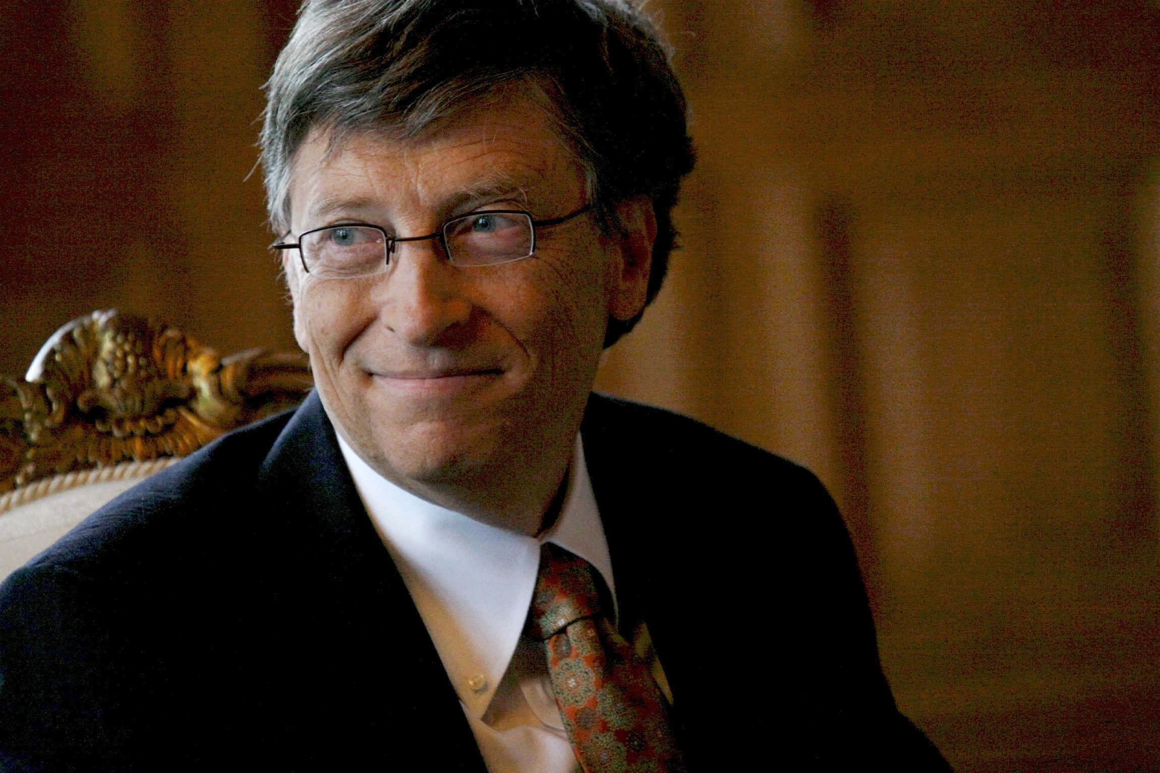 Bill Gates 桌布 壁紙 — 一位戴眼鏡微笑男士的特寫肖像，身穿深色西裝和圖案領帶，坐在華麗的椅子上。