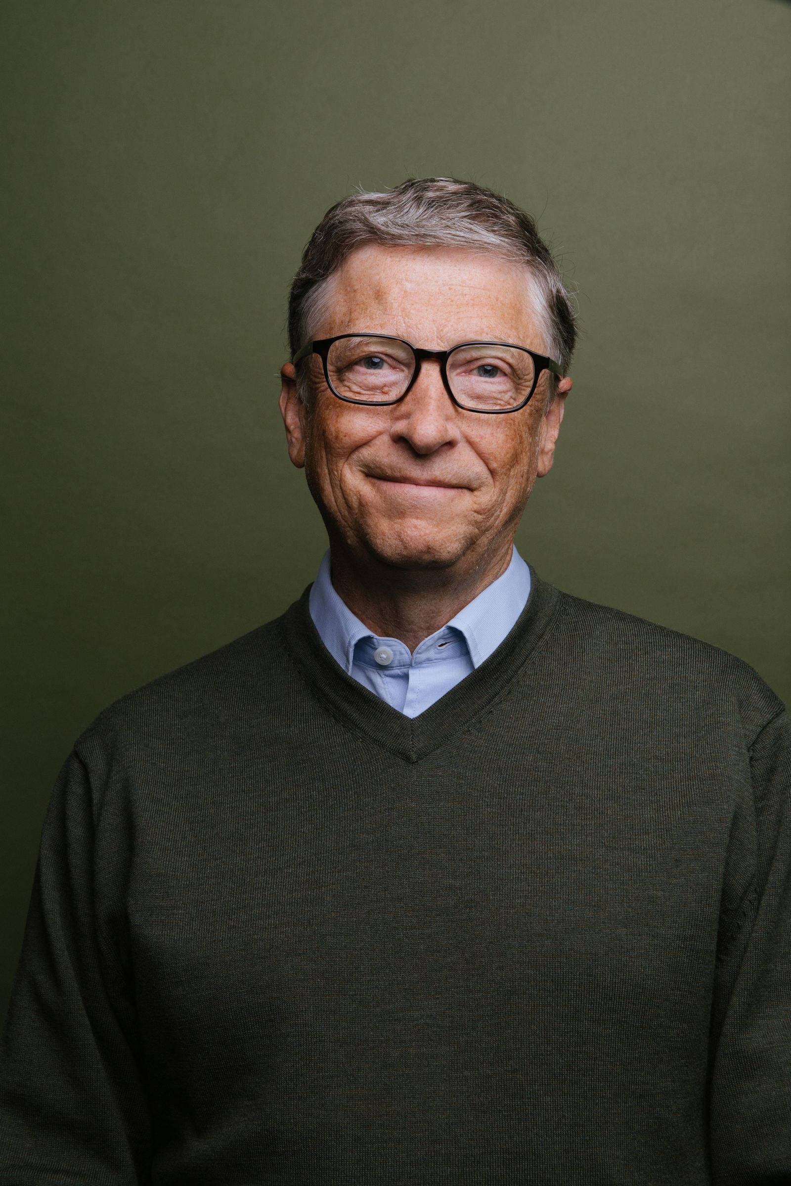 gates 桌布 壁紙 — 一位戴眼鏡微笑的年長男士的特寫肖像，身穿深綠色毛衣，內搭淺藍色有領襯衫，背景為純橄欖綠色。