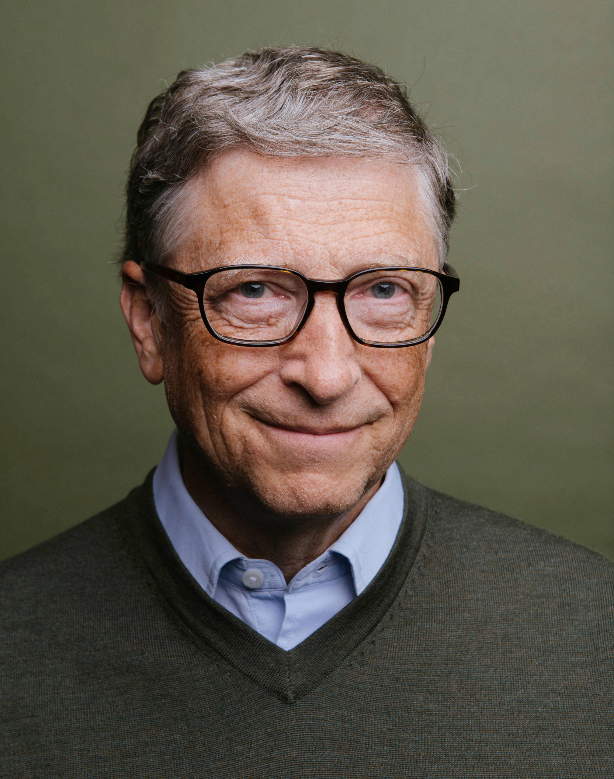 gates 桌布 壁紙 — 一位戴眼鏡微笑的年長男性特寫肖像，身穿綠色毛衣內搭淺藍色襯衫，背景為柔和橄欖色。