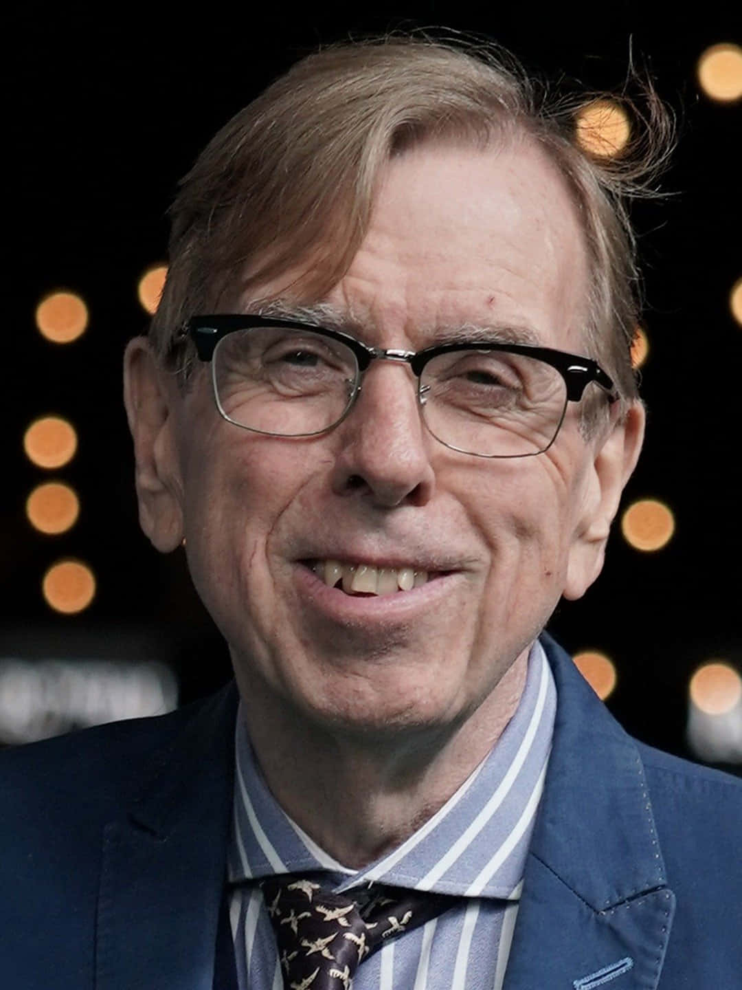 Timothy Spall 桌布 壁紙 — 一位戴眼鏡微笑的老年男士，身穿藍色西裝和圖案領帶，背景為深色帶有溫暖散景燈光。