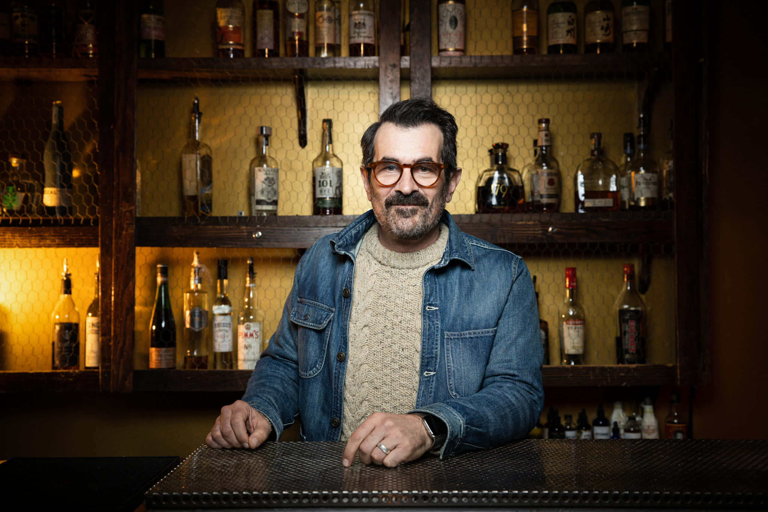 Ty Burrell 桌布 壁紙 — 一位戴眼鏡留鬍鬚的男人坐在吧檯前，身後有暖色燈光照射的酒瓶架。