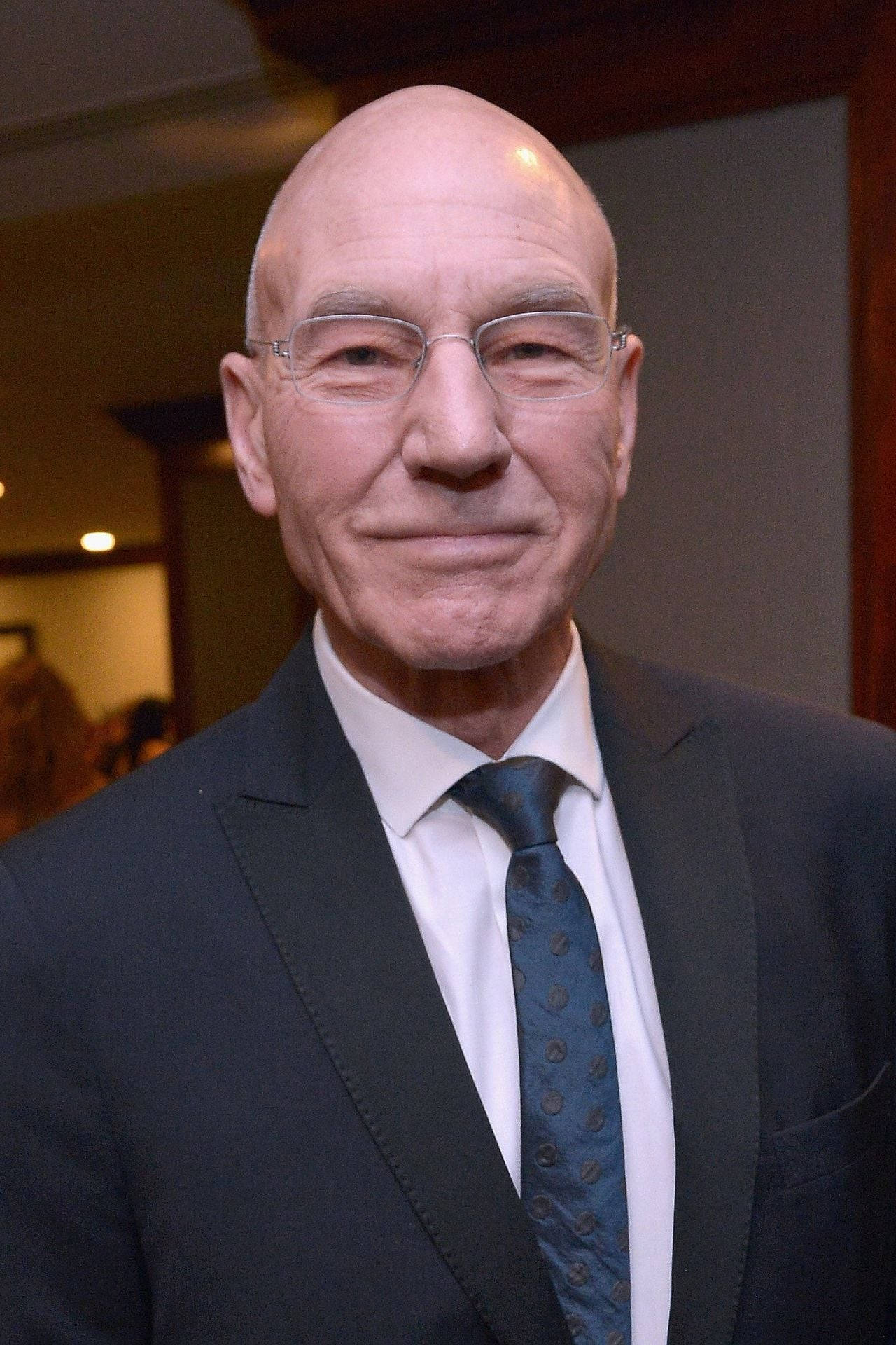 Celebrity 桌布 壁紙 — 一位戴眼鏡的光頭男士穿著深色西裝和圖案領帶的室內特寫肖像。