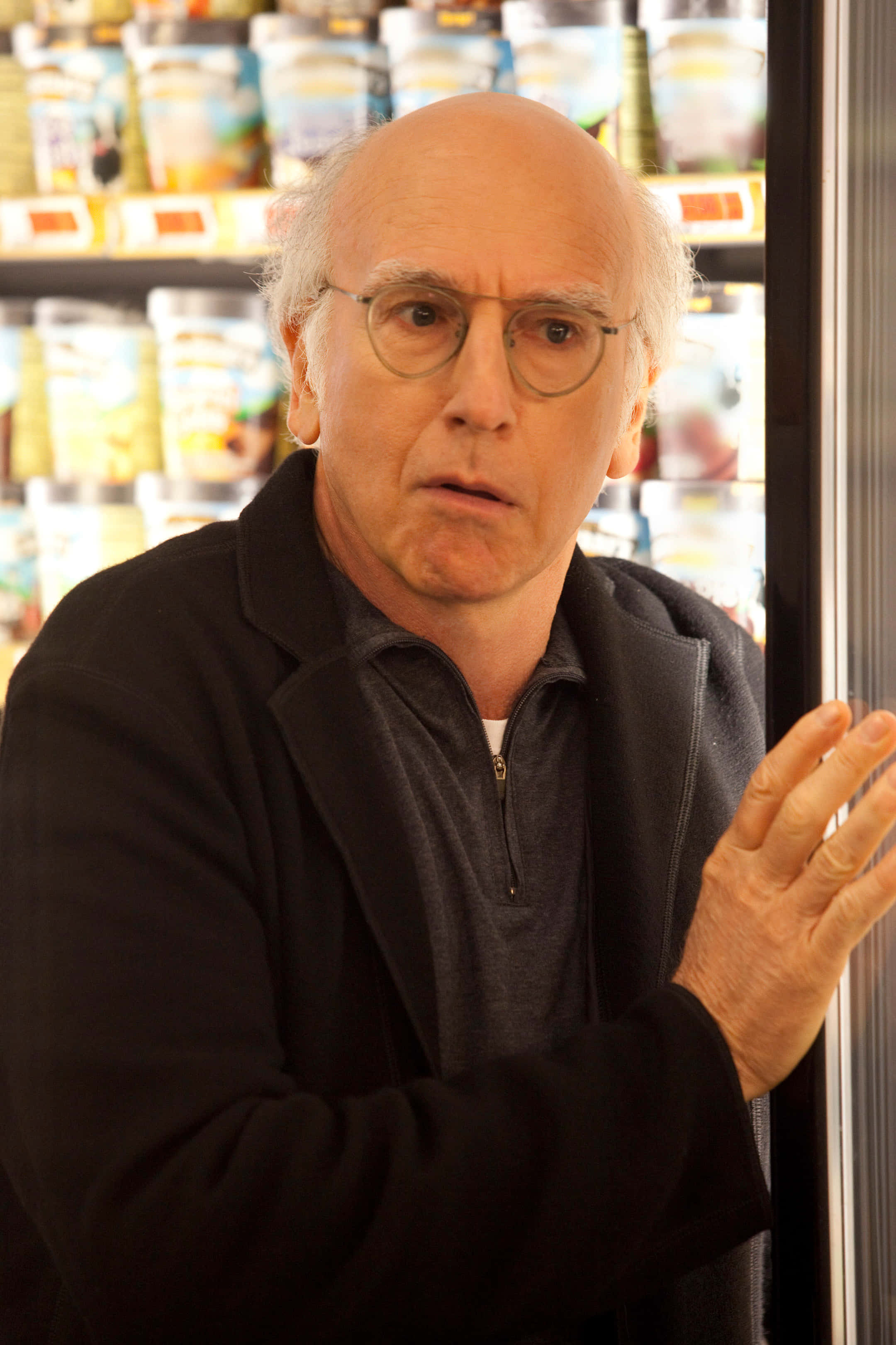 larry david 桌布 壁紙 — 一位戴眼鏡的光頭男子站在雜貨店走道中，表情驚訝。