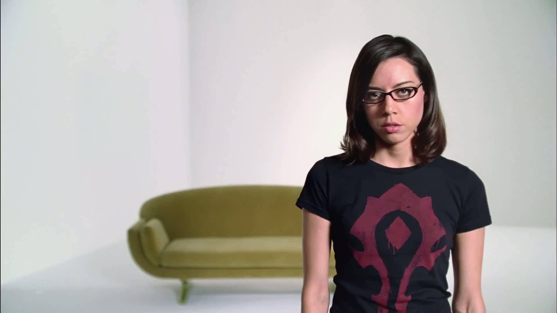 Aubrey 桌布 壁紙 — 一位戴眼鏡的女性，在一個簡約的白色房間裡，穿著一件帶有紅色遊戲標誌的黑色T恤，房間裡有一張綠色沙發。